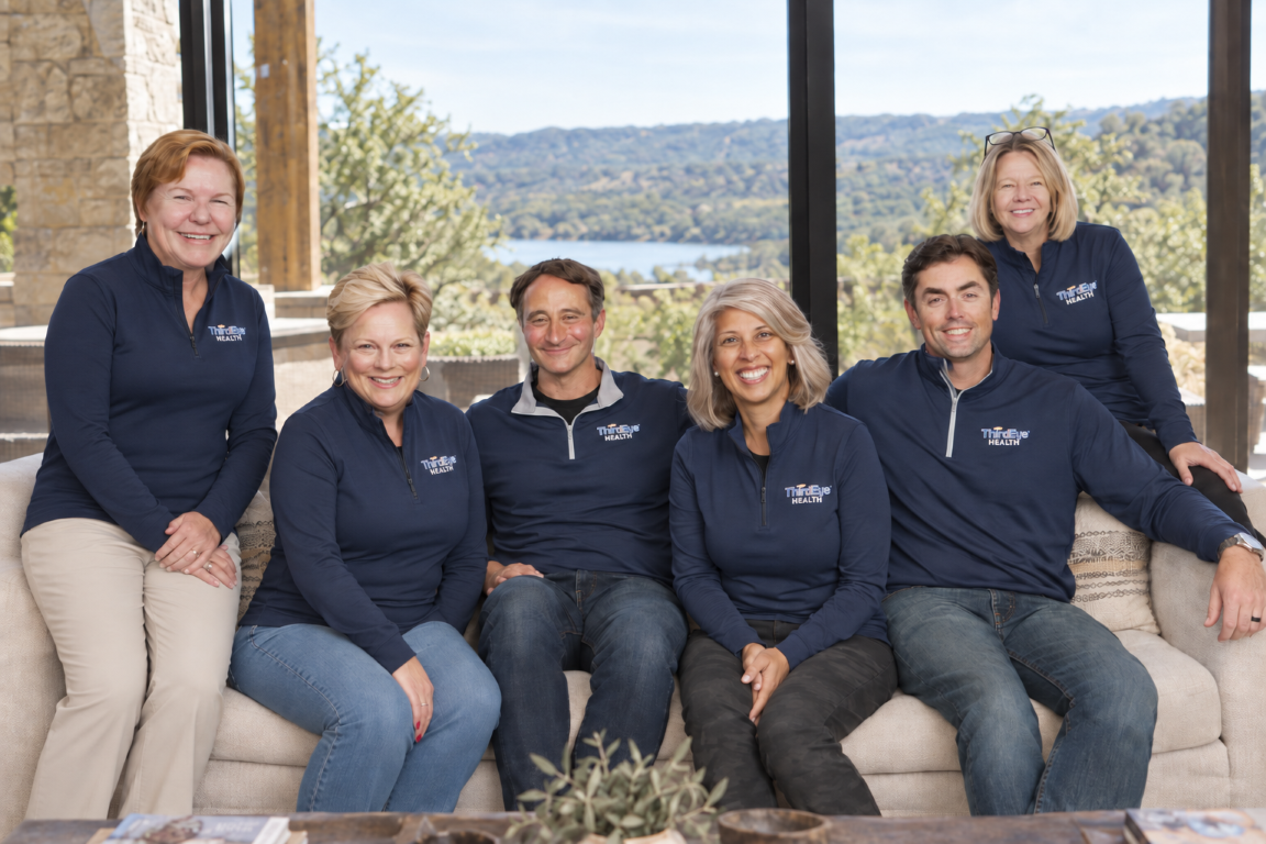 Six smiling adults sitting and standing on a beige couch in front of a large window with outdoor scenery, including trees, water, and hills. They are wearing matching navy blue jackets with a logo that says 'Third Eye Health'.