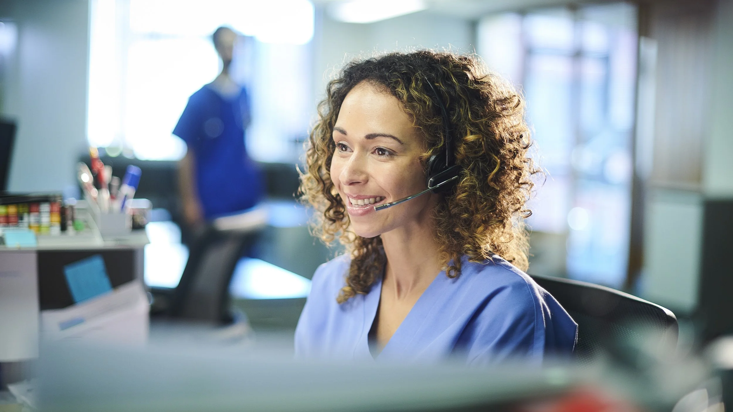 A smiling female nurse with curly hair wearing a headset and blue scrubs, working at a desk in a hospital or clinic.