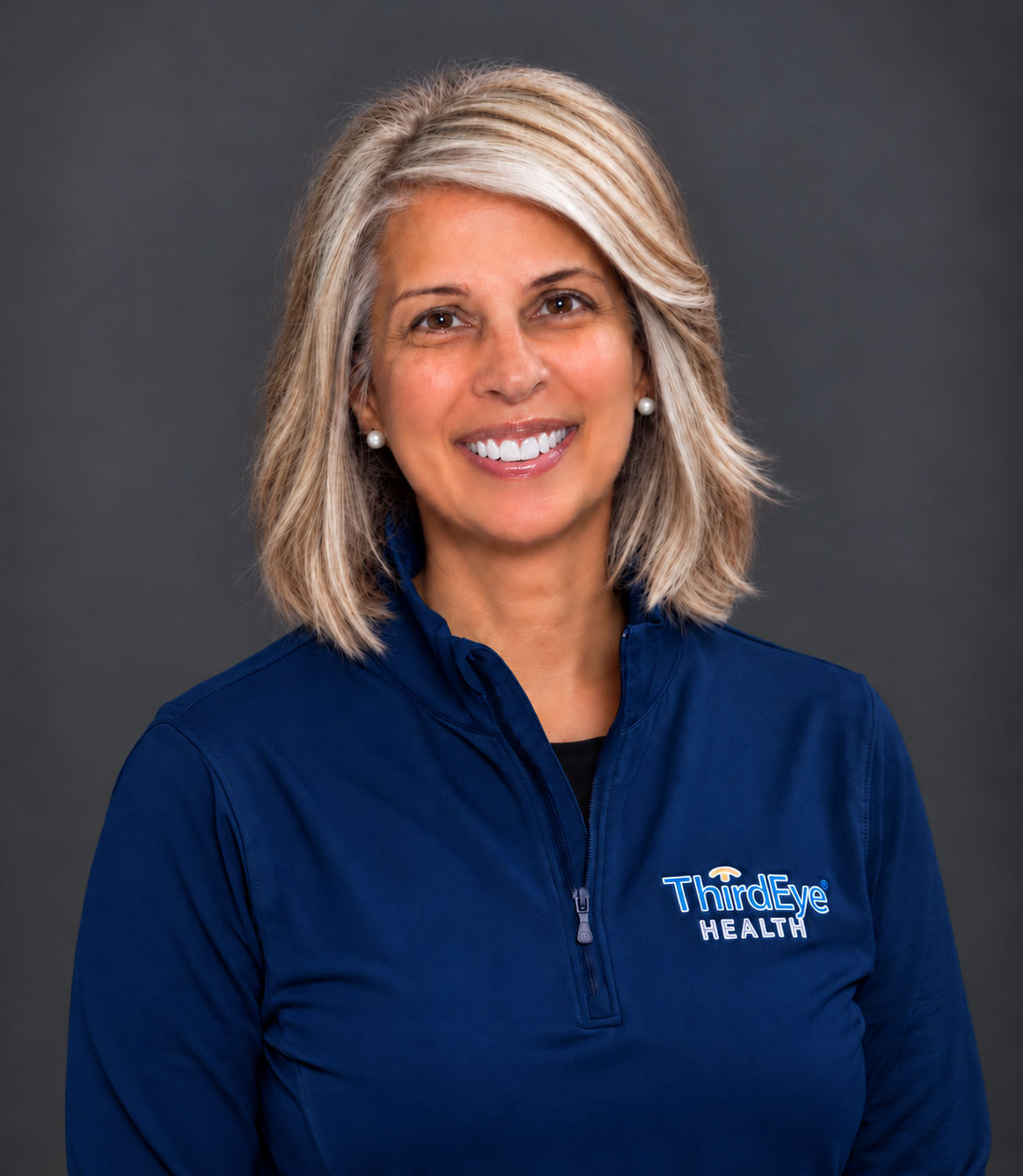 A smiling woman with blonde hair wearing a navy blue jacket with 'Third Eye Health' logo, pearl earrings, and a black shirt underneath, against a dark grey background.