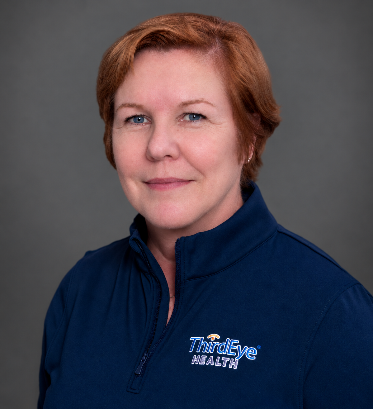 Portrait of a woman with short red hair and blue eyes, wearing a navy blue athletic jacket with the Third Eye Health logo, against a gray background.