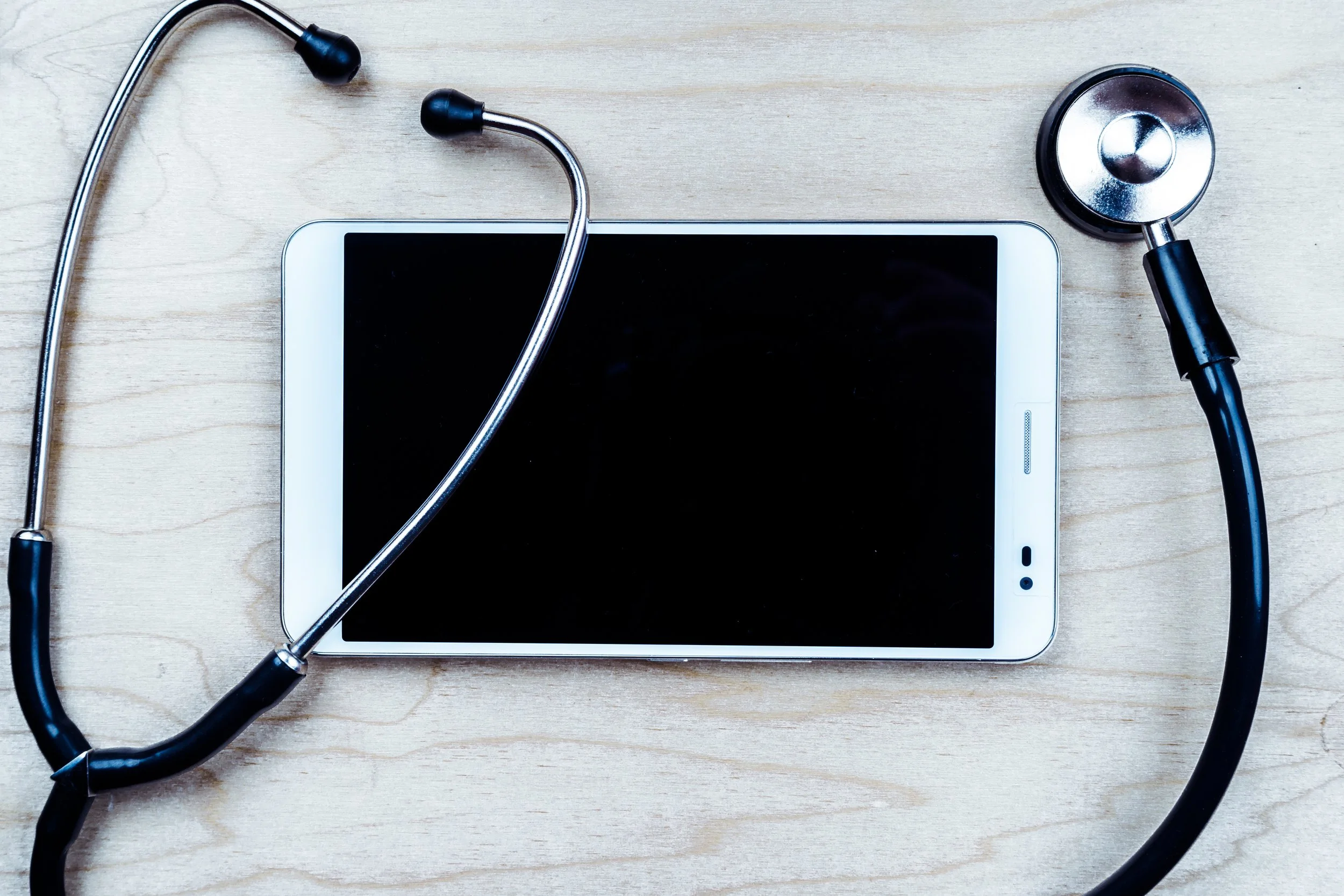 A stethoscope and a tablet on a light wooden surface.