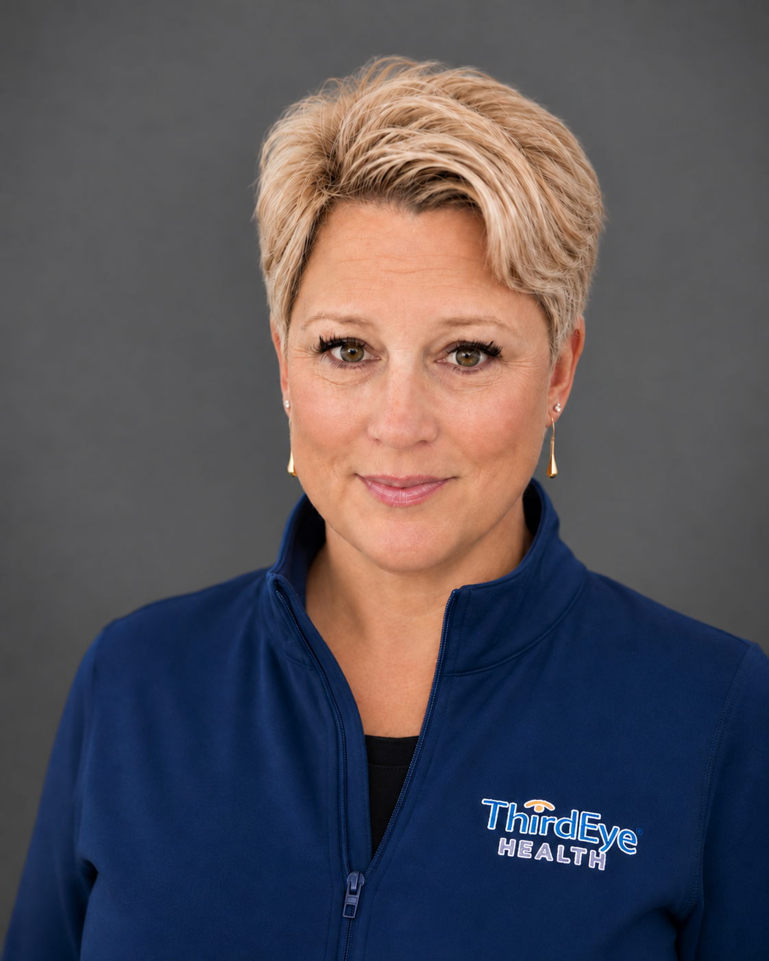 A woman with short blonde hair wearing a navy blue zip-up jacket with 'ThirdEye Health' embroidered on it, standing against a gray background.