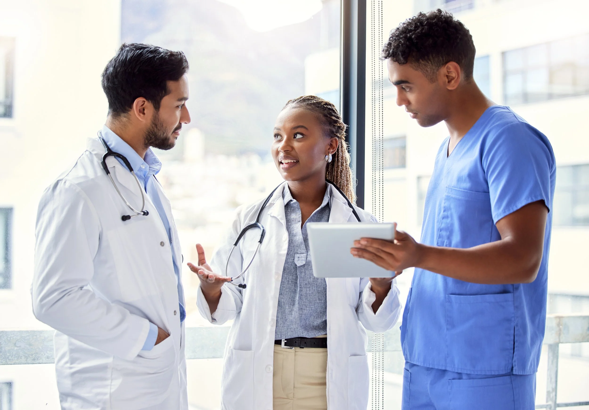 Three medical professionals, two men and one woman, are engaged in a discussion in a hospital or clinic. They are wearing medical scrubs and have stethoscopes around their necks. They are standing near a large window with a cityscape visible outside.