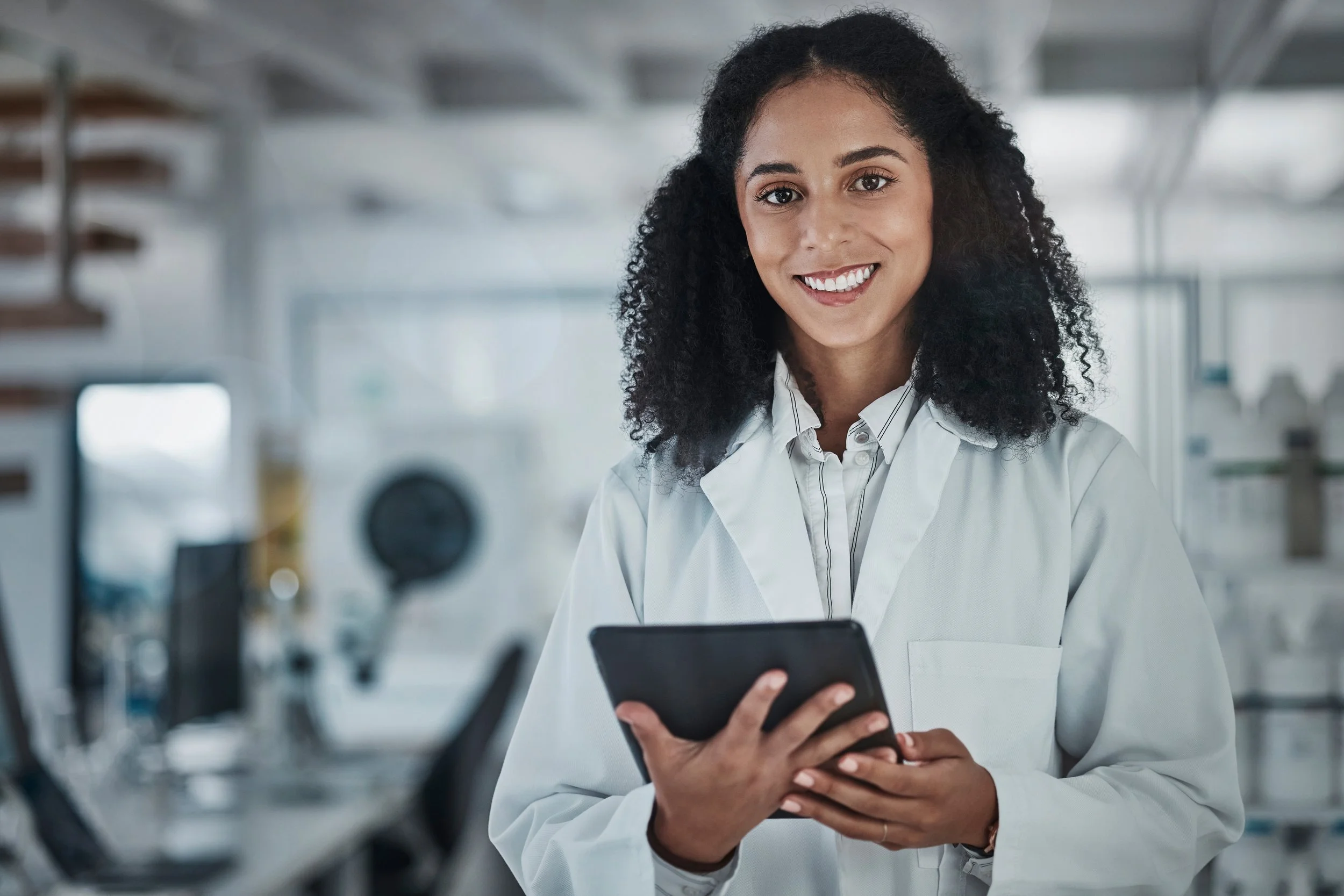 A woman in a white lab coat smiling and looking at her phone in a laboratory setting.