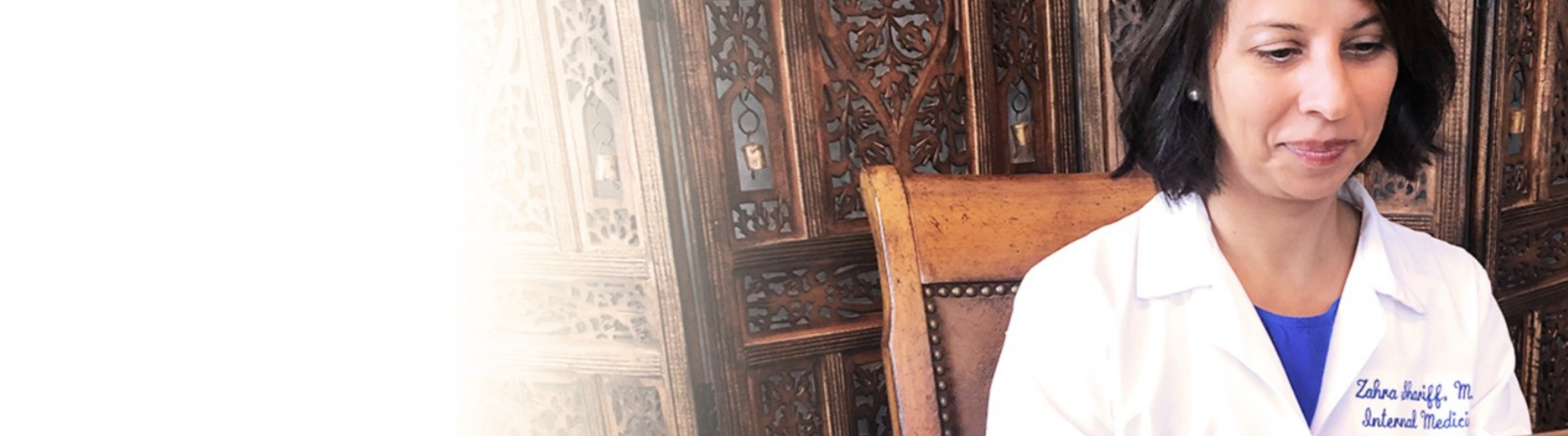 A woman with dark hair wearing a white coat with blue embroidery, sitting on a wooden chair in front of a carved wood screen.