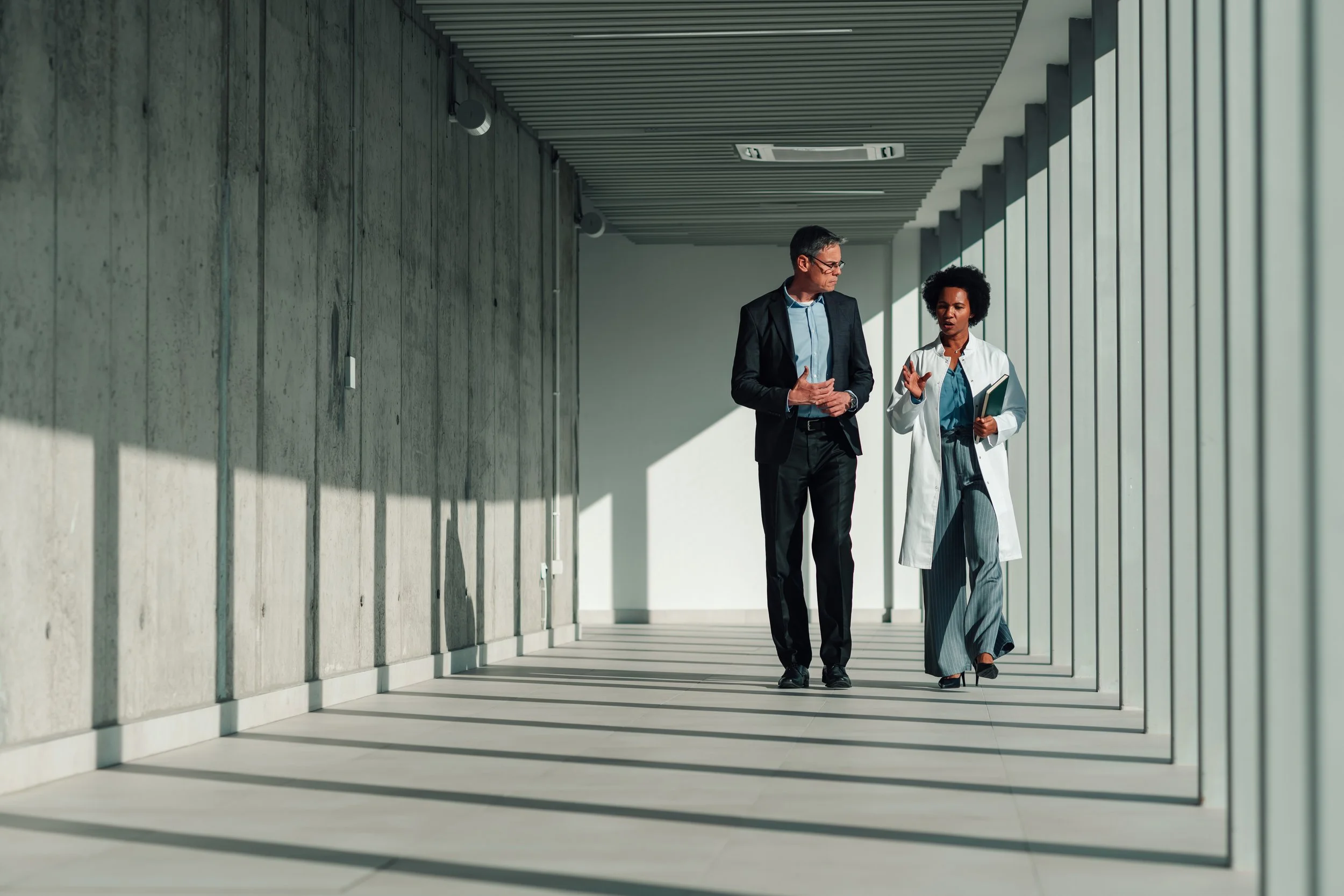 A man and a woman walking and talking in a modern corridor with concrete walls and large windows. The man is dressed in a black suit and glasses, and the woman is wearing a white coat and holding a tablet.