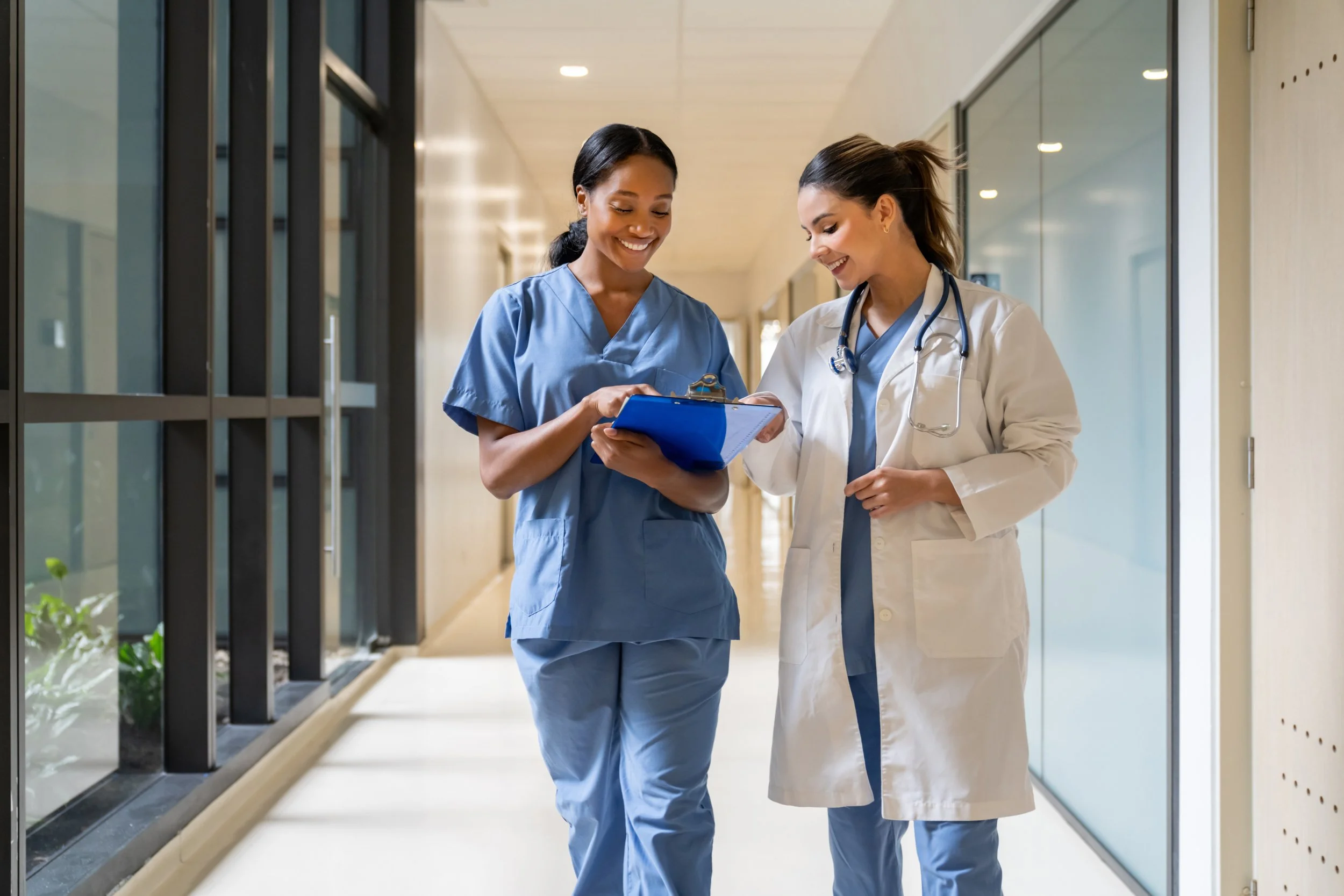 Two female healthcare professionals, one in blue scrubs and the other in white lab coat with a stethoscope, smiling and looking at a clipboard while standing in a hospital hallway.