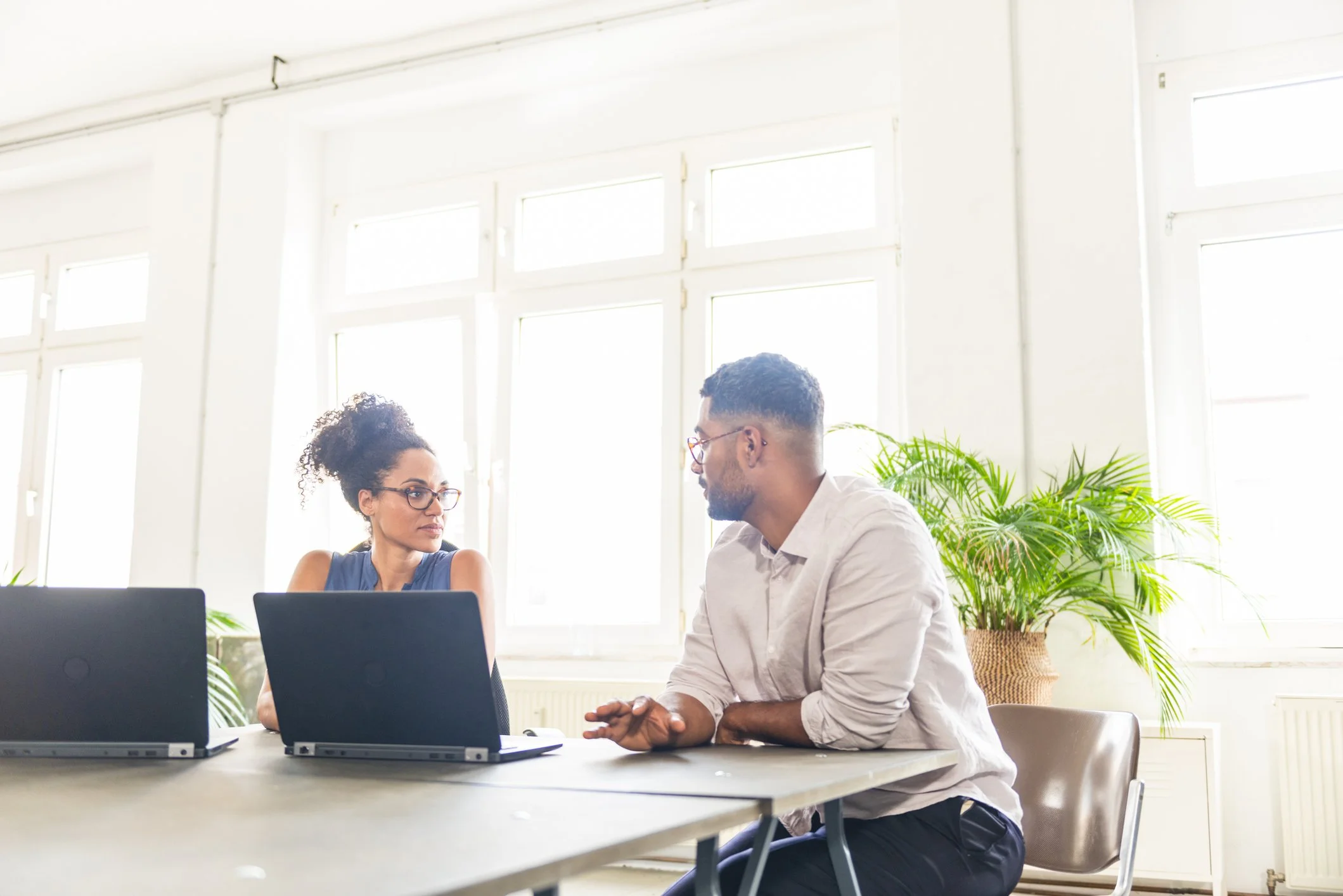 Two people engaged in a conversation at a desk in a well-lit office with large windows and green potted plants.