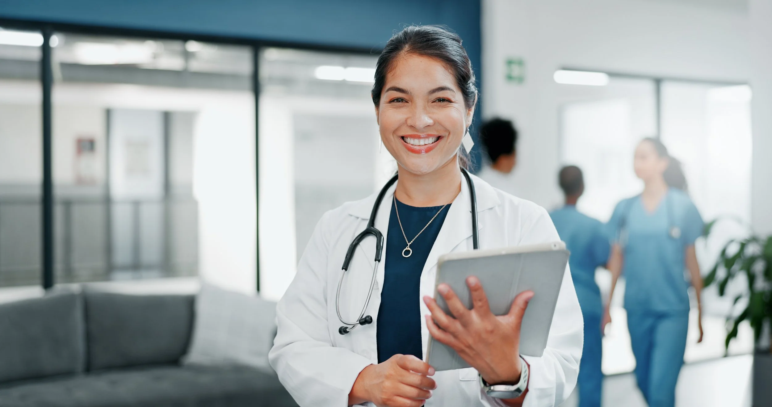 A smiling female doctor holding a tablet in a hospital lobby with other medical staff in the background.