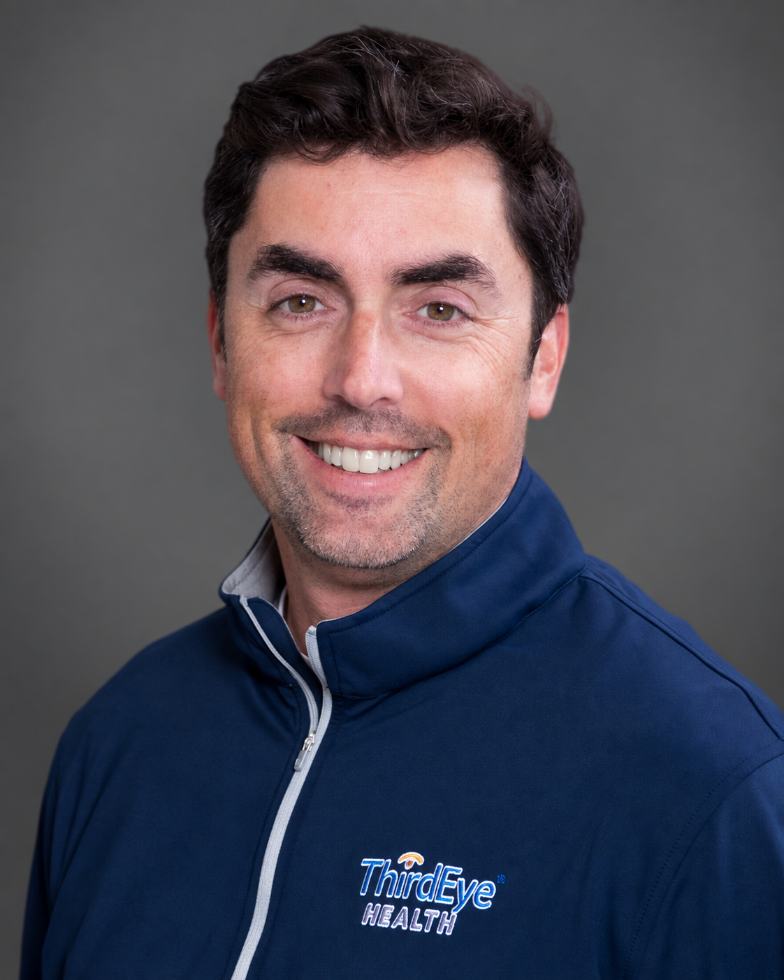 Headshot of a smiling man with brown hair wearing a navy blue zip-up shirt with Third Eye Health logo on it, against a neutral background.