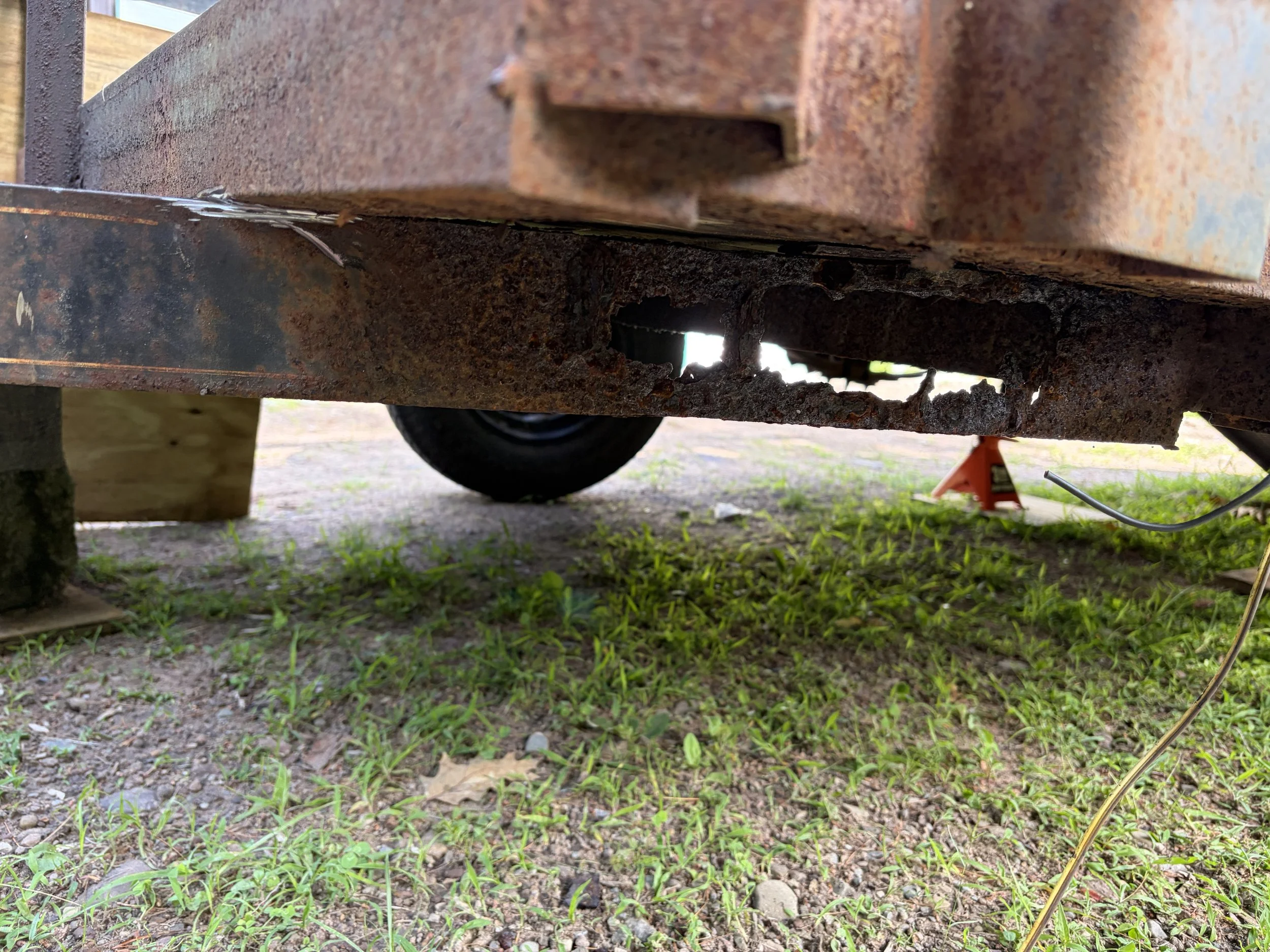 Close-up of a rusty, corroded metal structure or piece of scrap metal outdoors, with grass and dirt underneath and a wheel in the background.