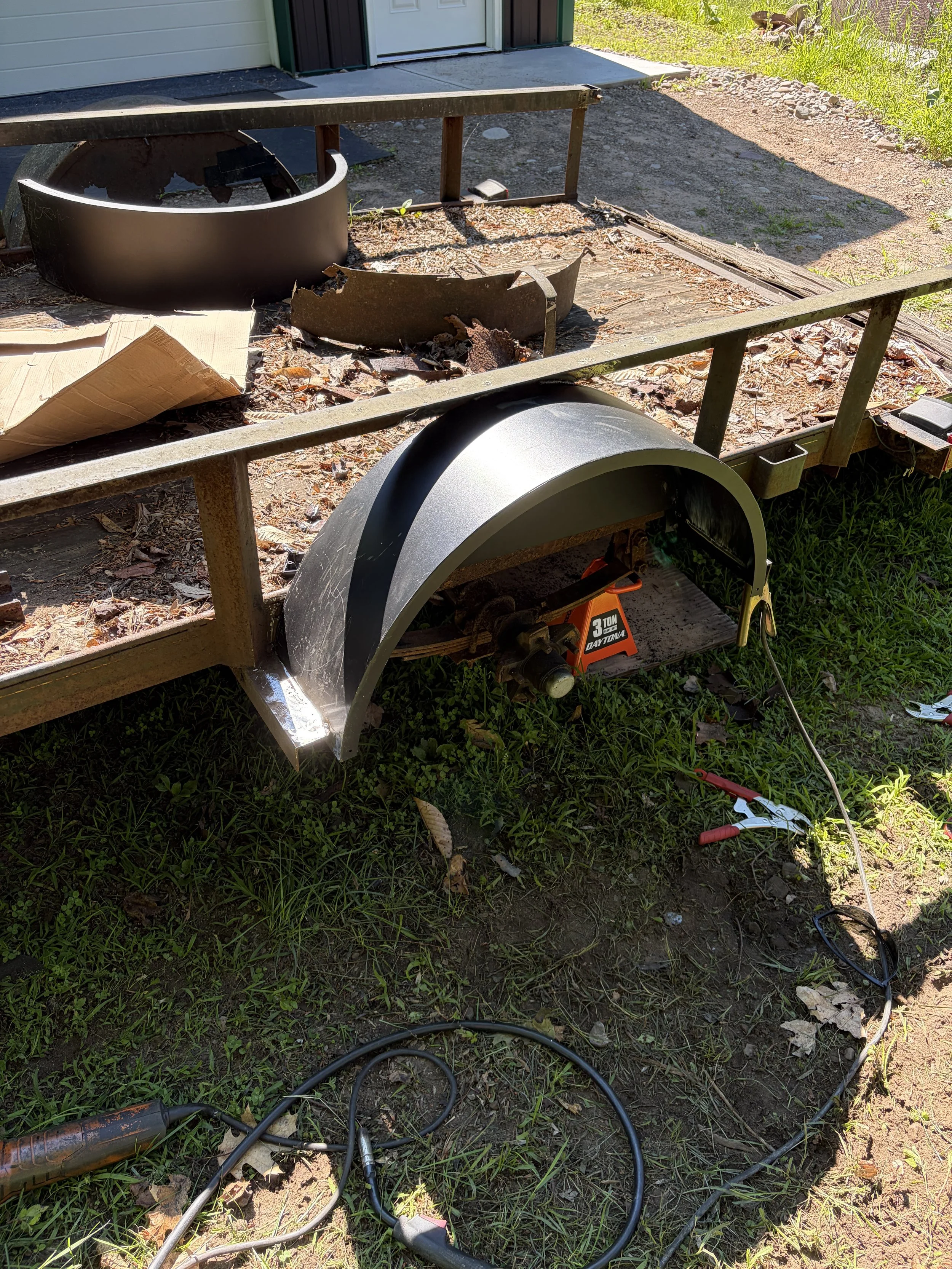 A rusty work trailer with a black grill and welding equipment lying on the ground, outside a building with a gray door. The trailer is parked on grass and dirt, with power cables and tools scattered around.