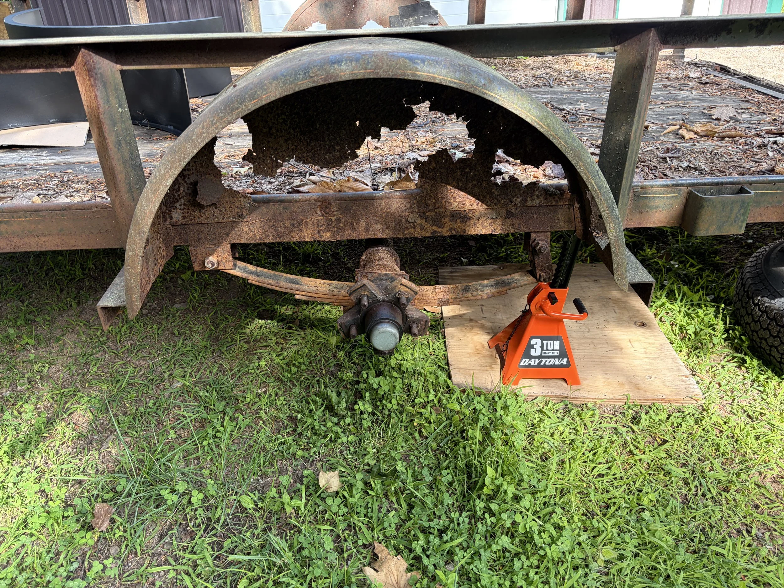 Rusty metal roller on a trailer, surrounded by leaves and grass in an outdoor yard, with a 3-ton heavy-duty Dayton jack stand supporting the trailer on wood.