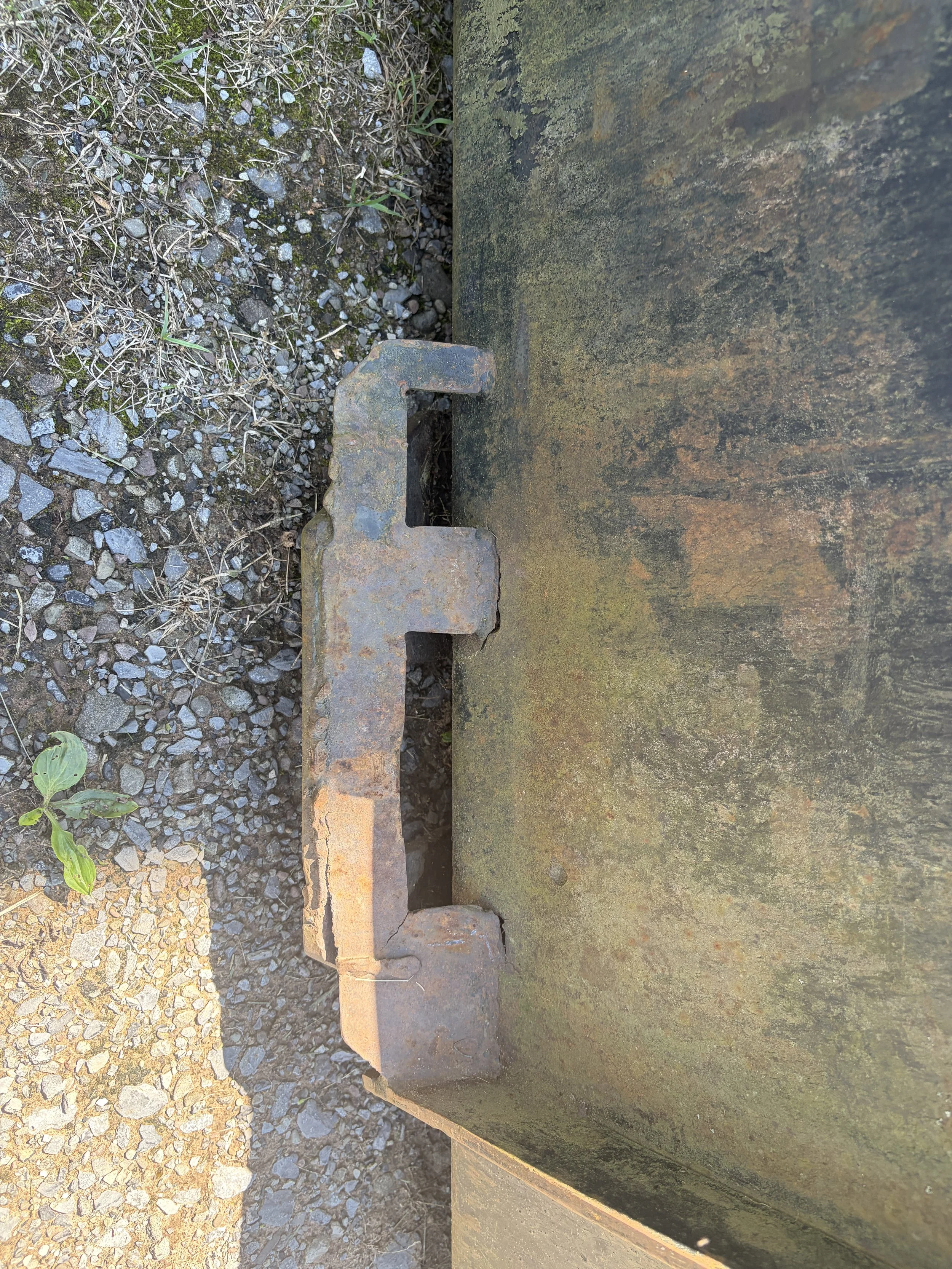 Close-up of a rusty metal hook attached to a weathered, moss-covered concrete wall, with gravel ground and some small green plants nearby.