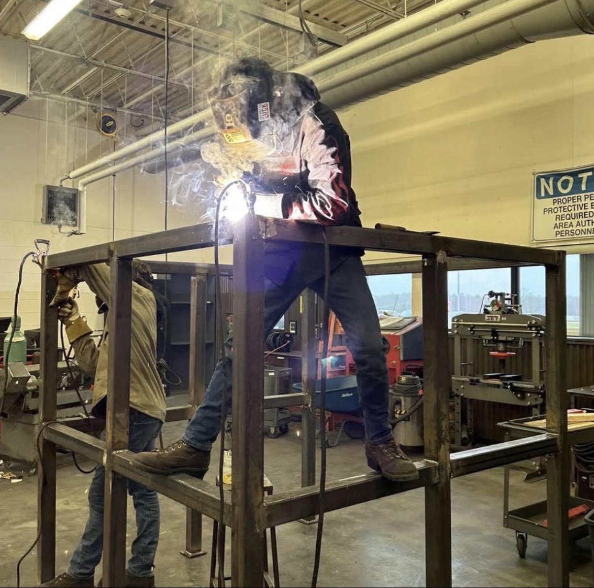 A person welding inside a metal frame structure in a workshop, with protective gear and sparks flying, while another worker stands nearby.