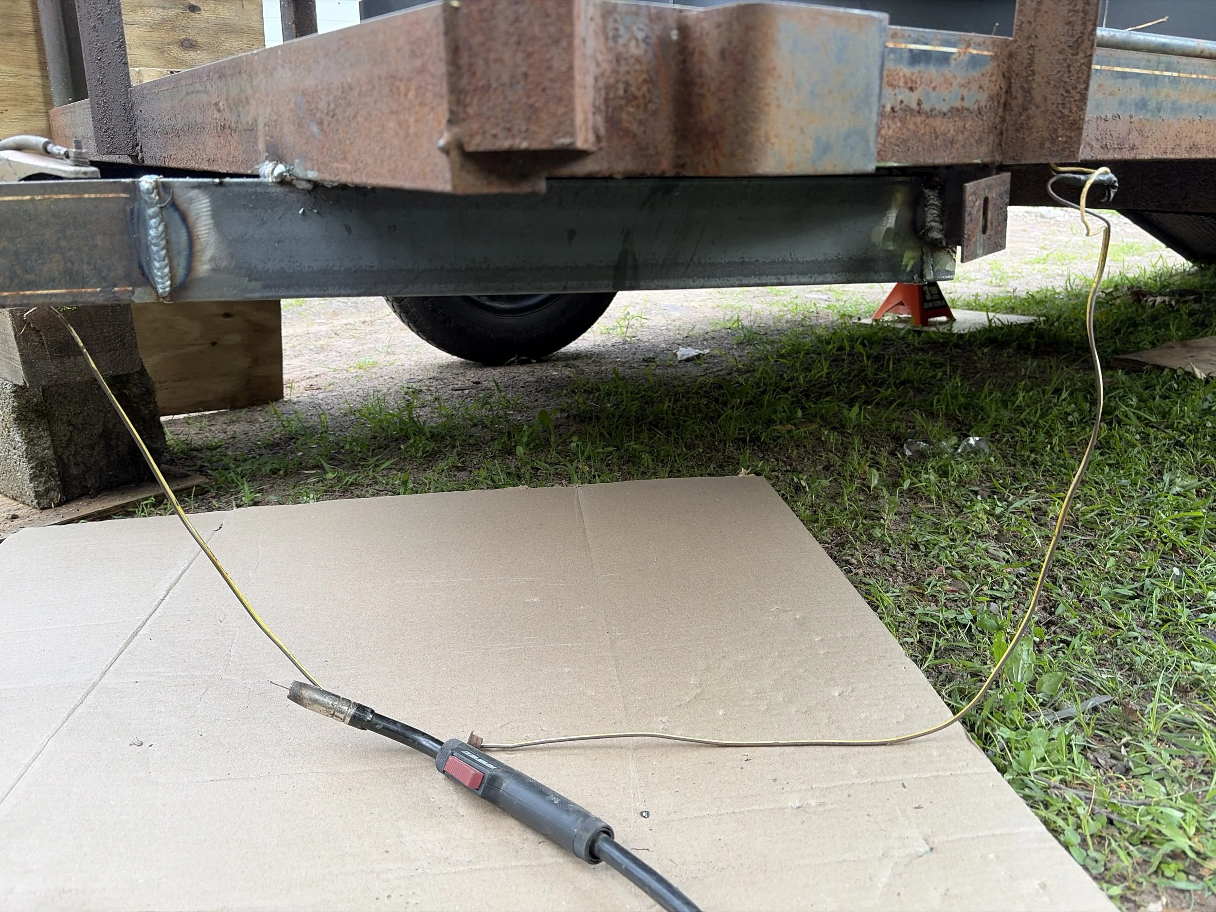An image showing a construction or repair scene with a metal trailer frame rusted on top of a black metal support. A welding tool with a power cord is lying on a piece of cardboard on the ground. There are some yellow strings and a jack stand with a label underneath the trailer. The scene appears outdoors with some grass and dirt visible.