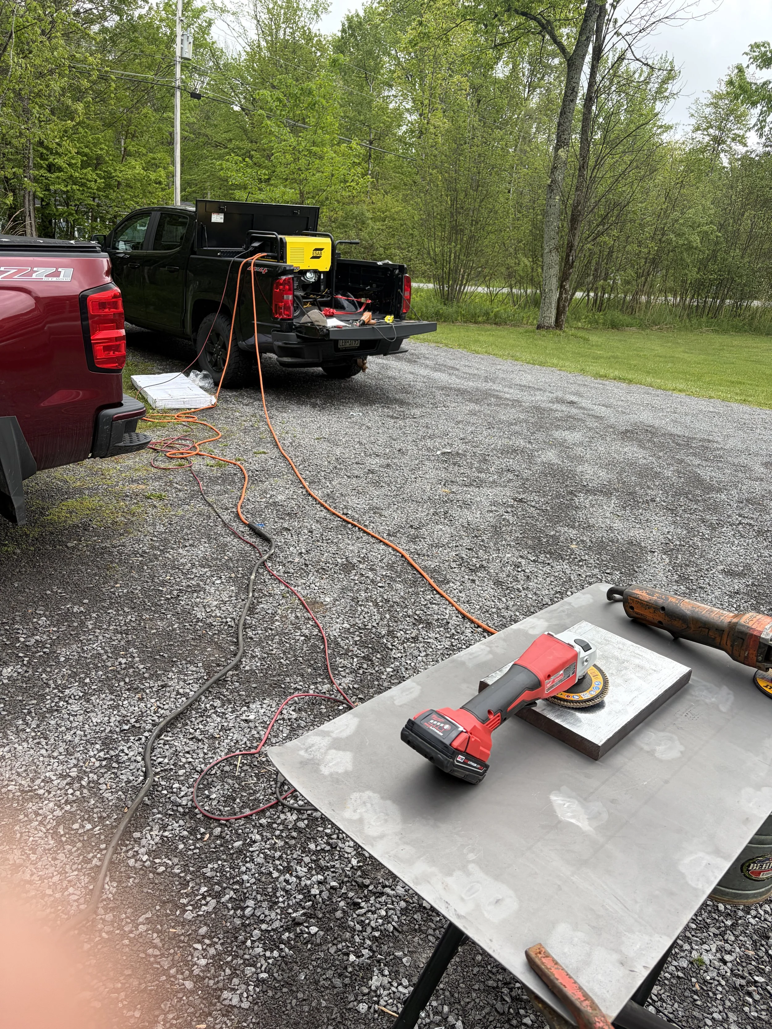 A gravel driveway with work tools and equipment including a cordless drill, angle grinder, and various cords, set on a metal table and on the ground. In the background, there are two trucks, one black and one red, with the bed of the black truck containing tools and equipment. Surrounding the area are trees and a grassy area.