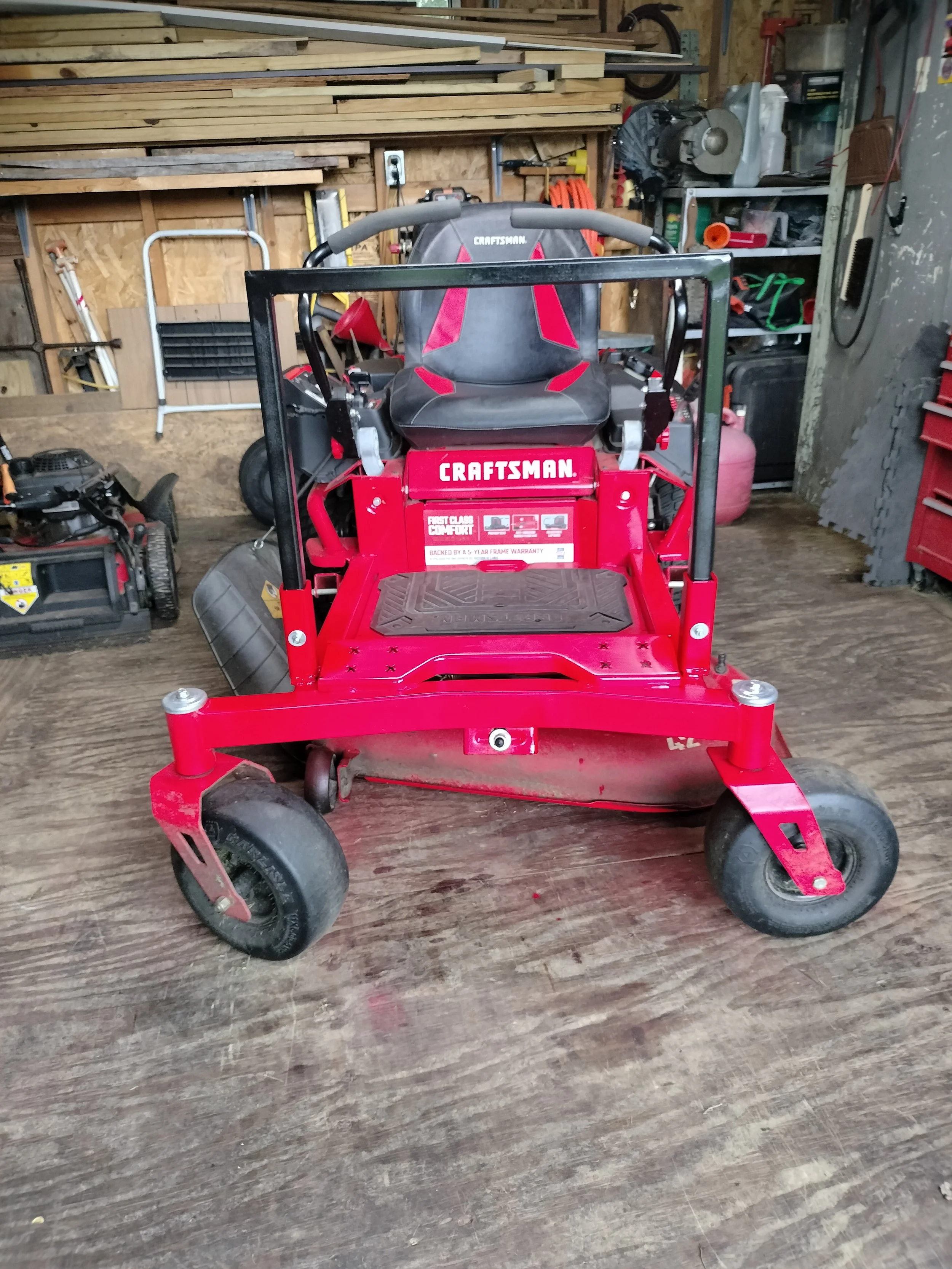 A red Craftsman lawnmower with a black seat and handles is in a wooden shed or workshop with shelves and tools in the background.