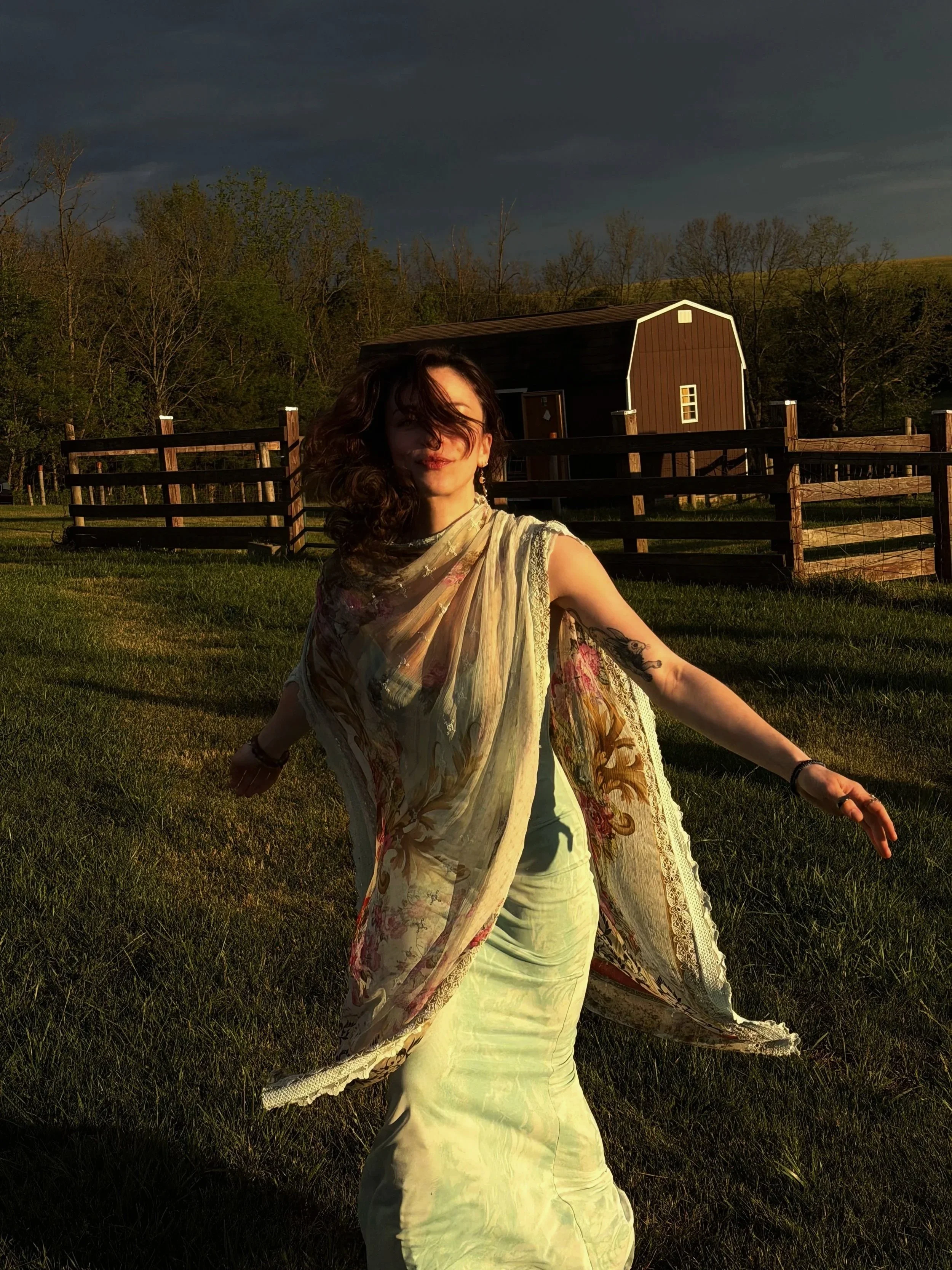 A woman dancing or posing in a field during sunset with a dark, cloudy sky and a red barn in the background.