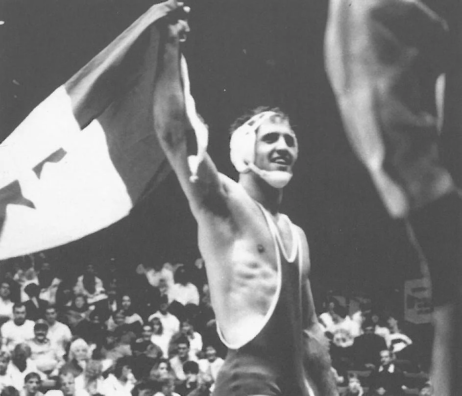 A male wrestler with headgear and singlet, smiling and holding a Canadian flag, standing in front of a crowd.