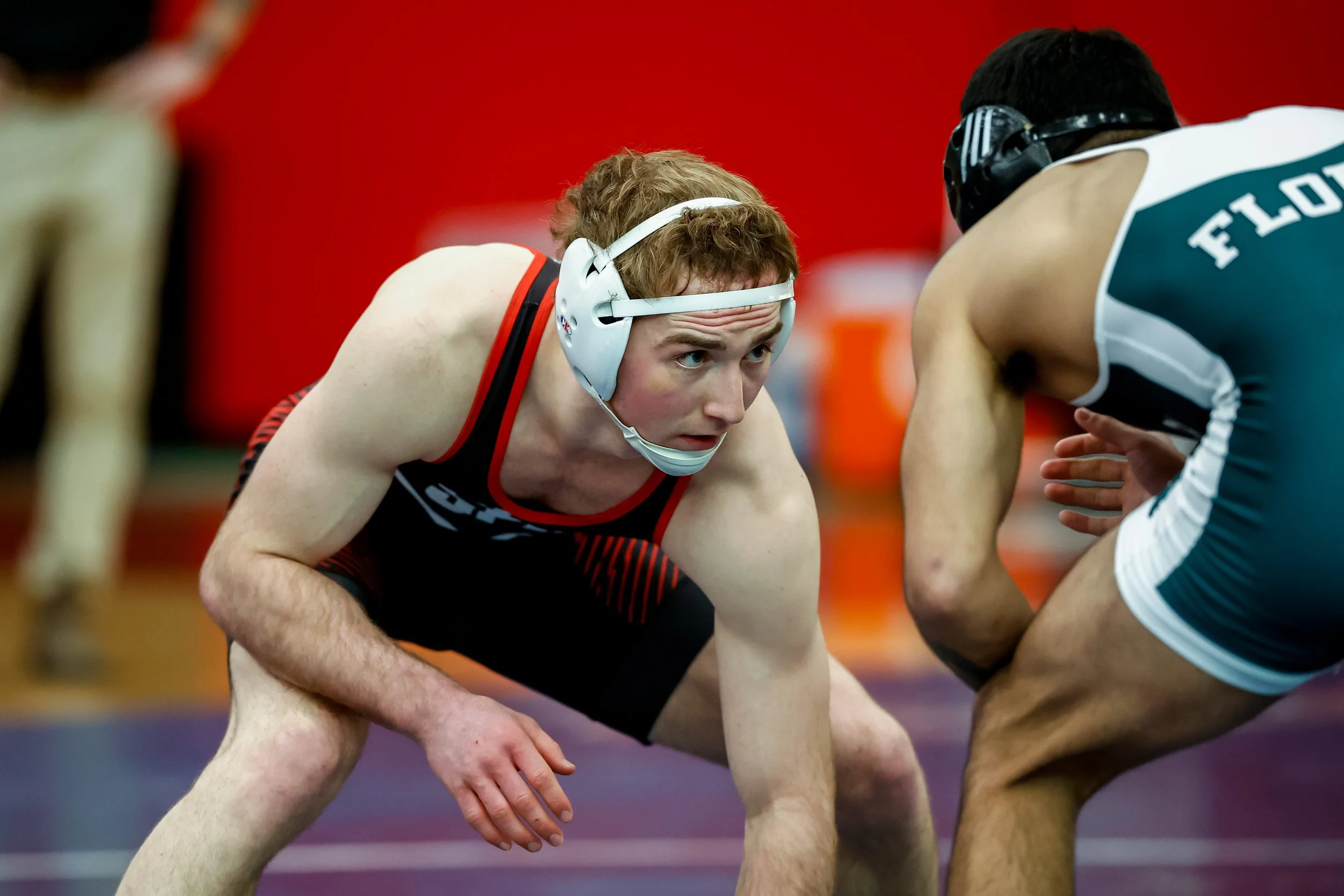 Two male wrestlers in a match, one in a red and black singlet and white headgear, and the other in a teal and white singlet, crouching and preparing for a move.