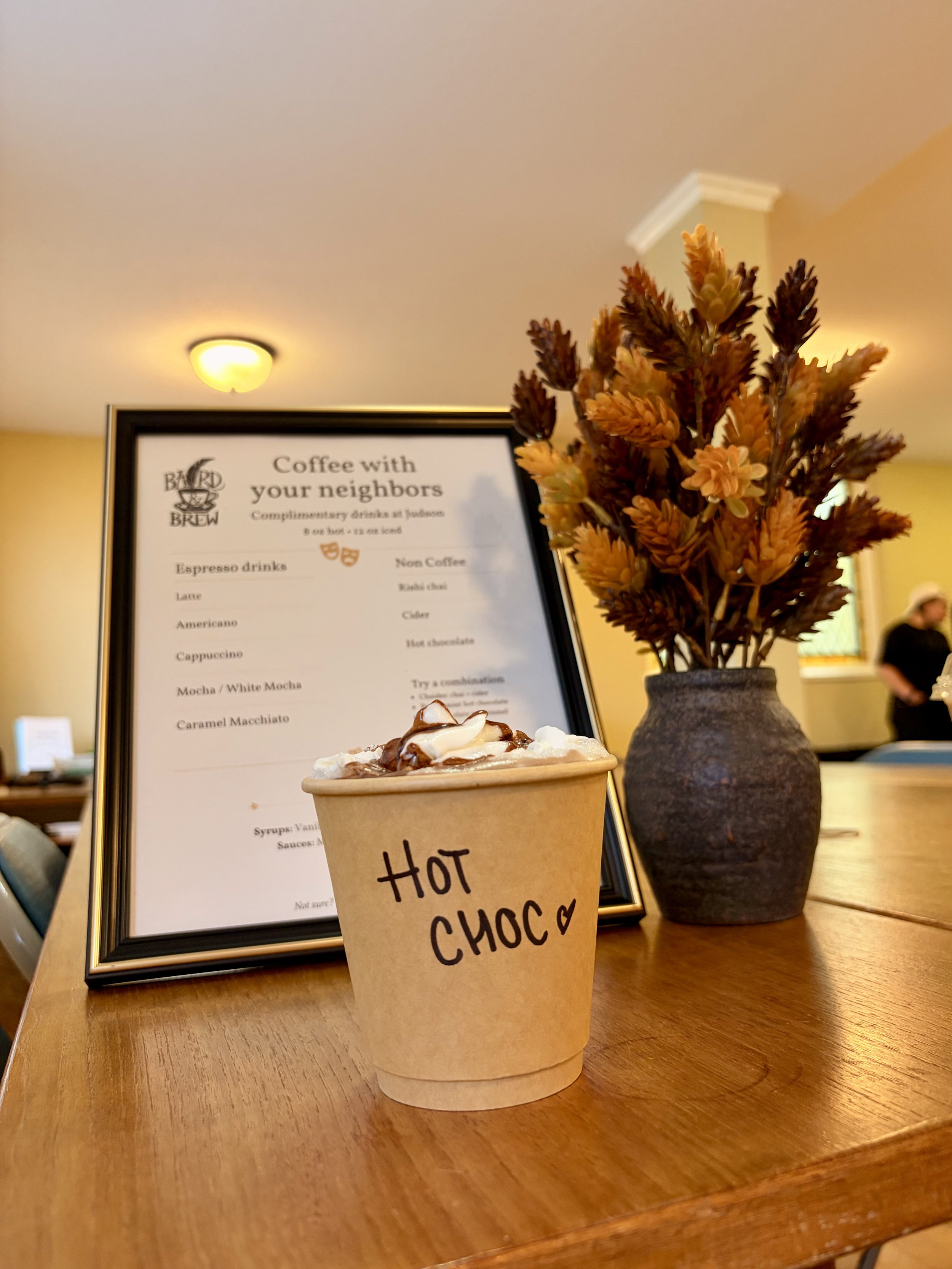 Bard & Brew hot chocolate topped with whipped cream and chocolate drizzle beside a menu and autumn flowers.