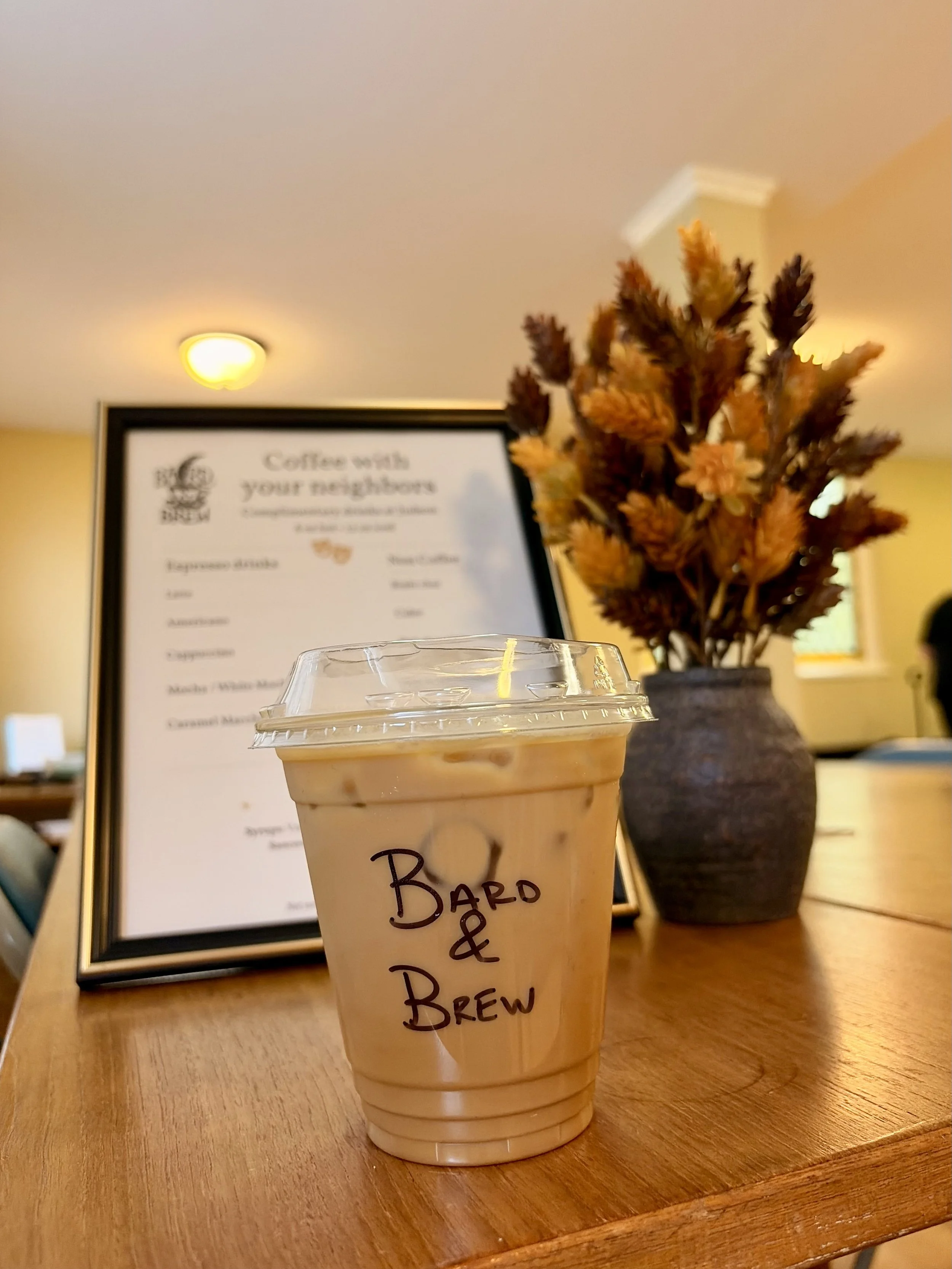 Bard & Brew iced latte beside a framed drink menu and autumn flowers.