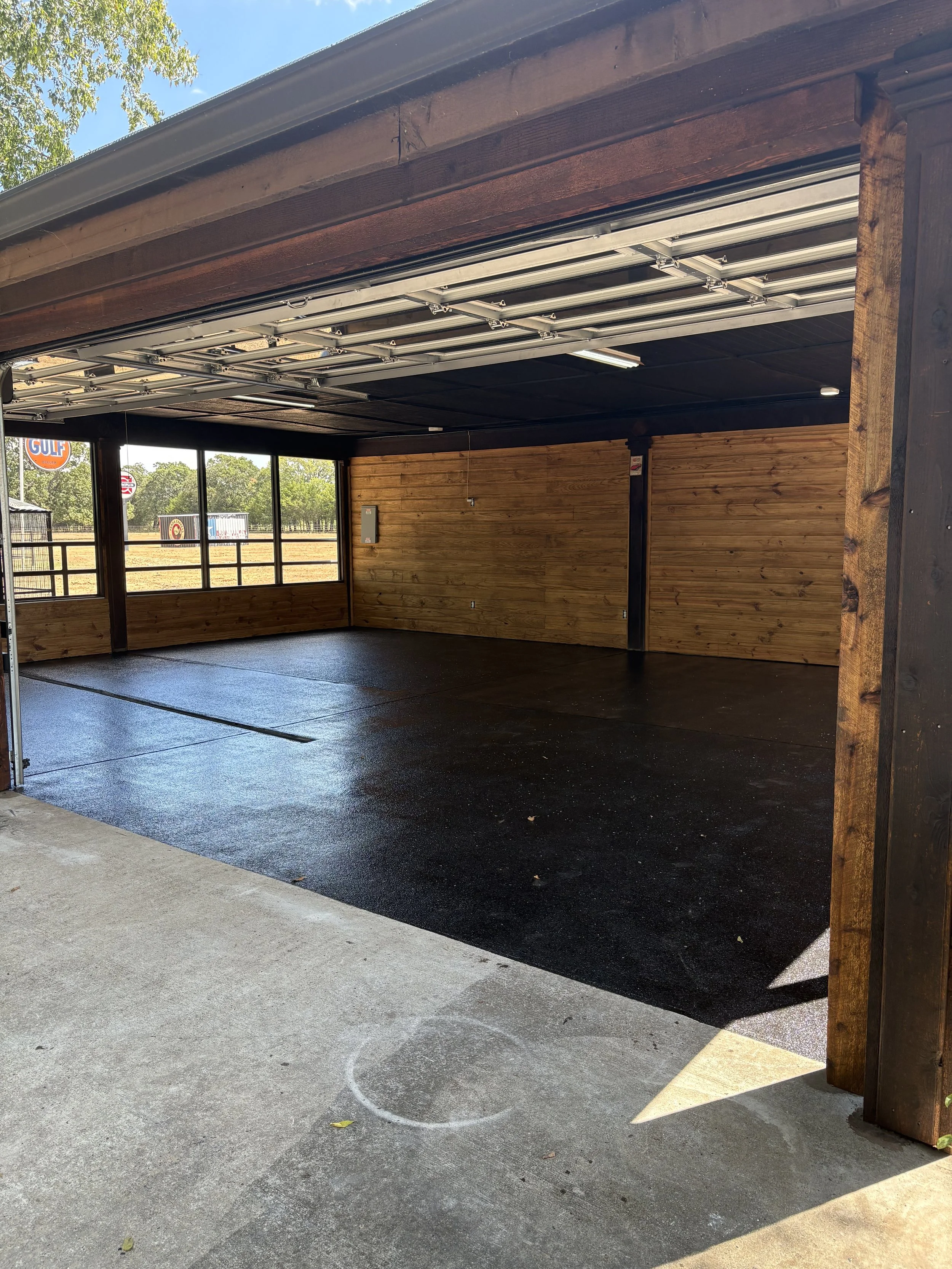 Covered outdoor structure with wood walls, a black floor, and a garage-style door that is partially open. Outside, there are trees and signs visible through windows.