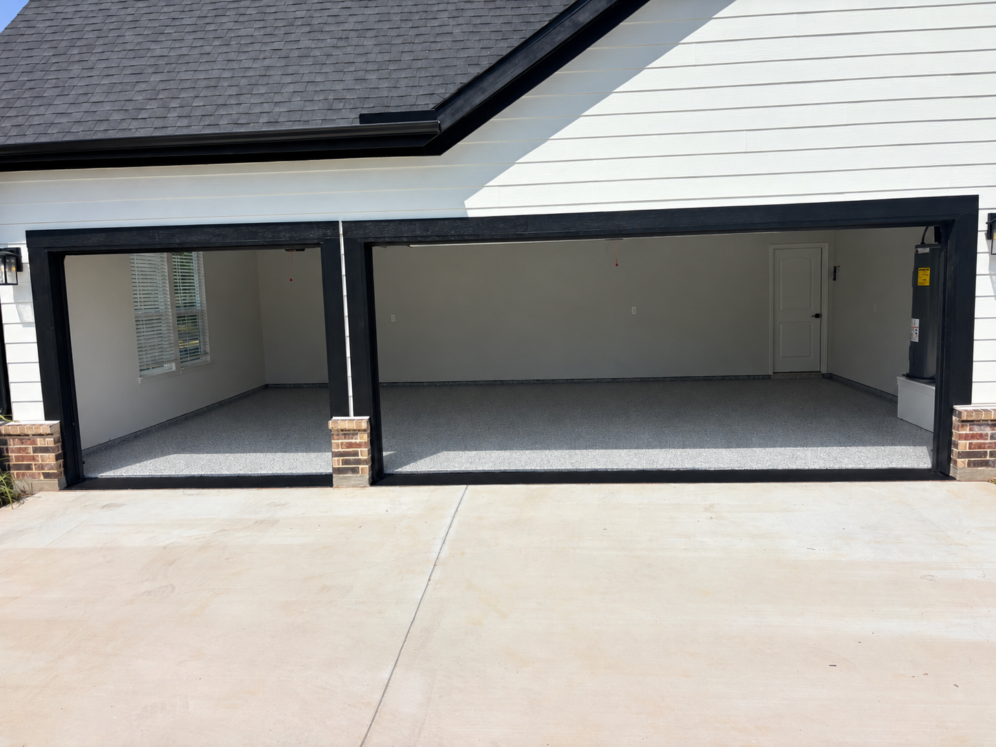 Empty two-car garage with gray flooring, white walls, and a white door at the back. The garage has black trim around the open doors and brick accents at the base. Outside, there is a concrete driveway.