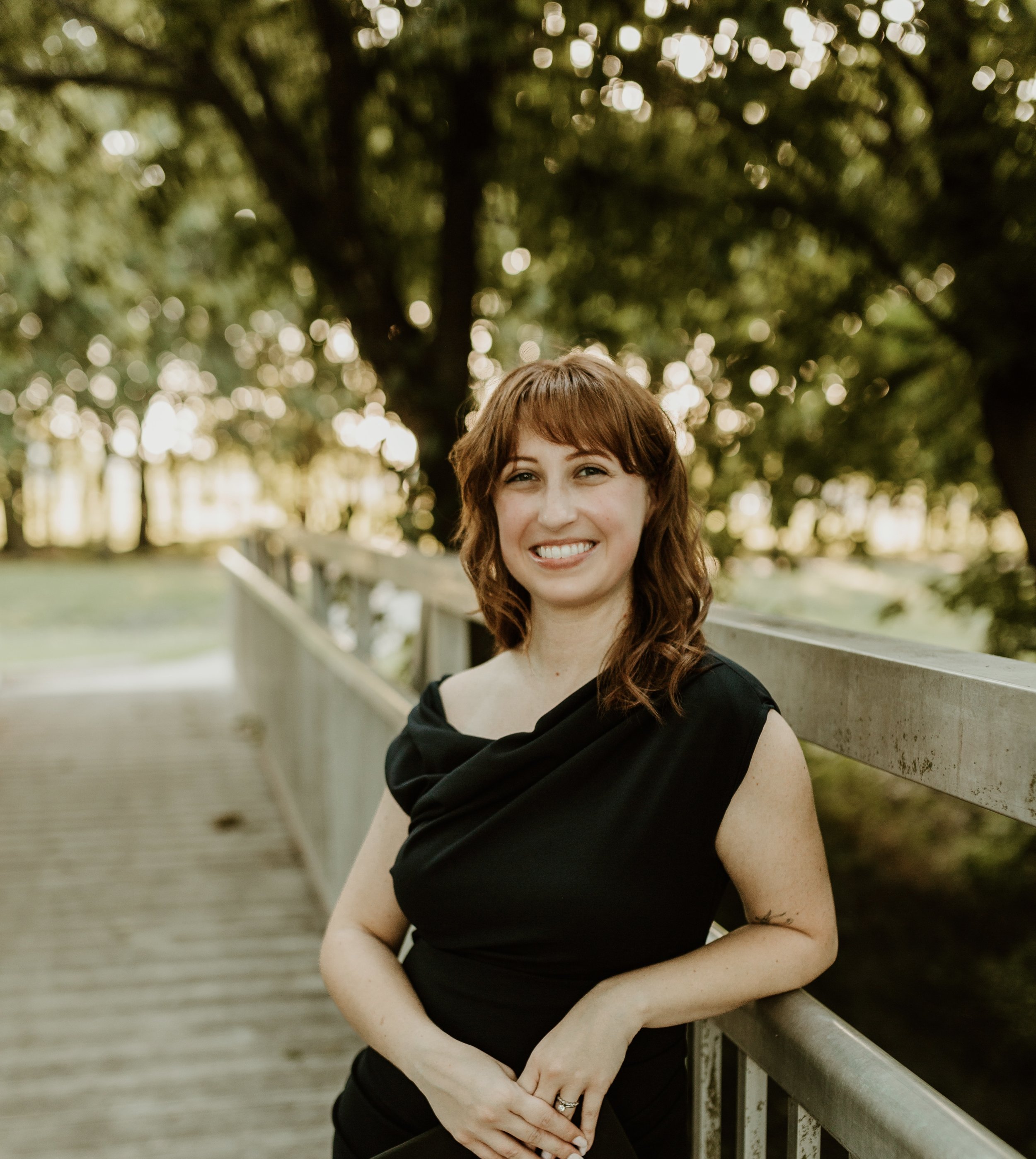 A smiling woman with reddish-brown hair, wearing a black dress with an off-shoulder design, stands outdoors on a wooden bridge with green trees and blurred lights in the background.
