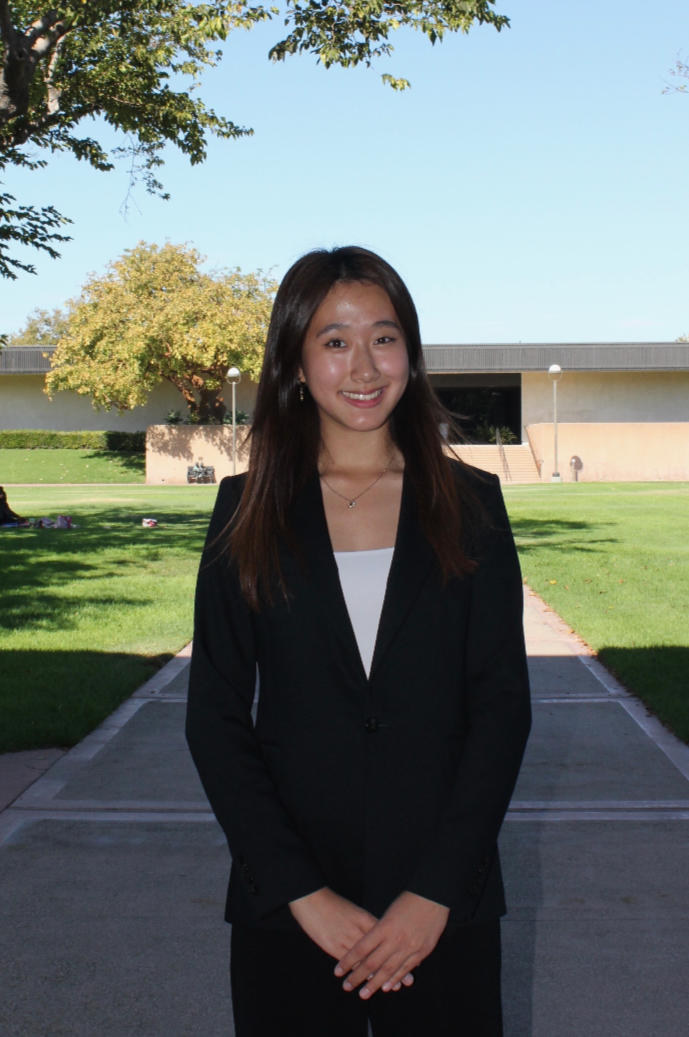 A young woman dressed in a black blazer and white top standing outdoors on a sunny day, with a school or campus building, trees, and a lawn in the background.