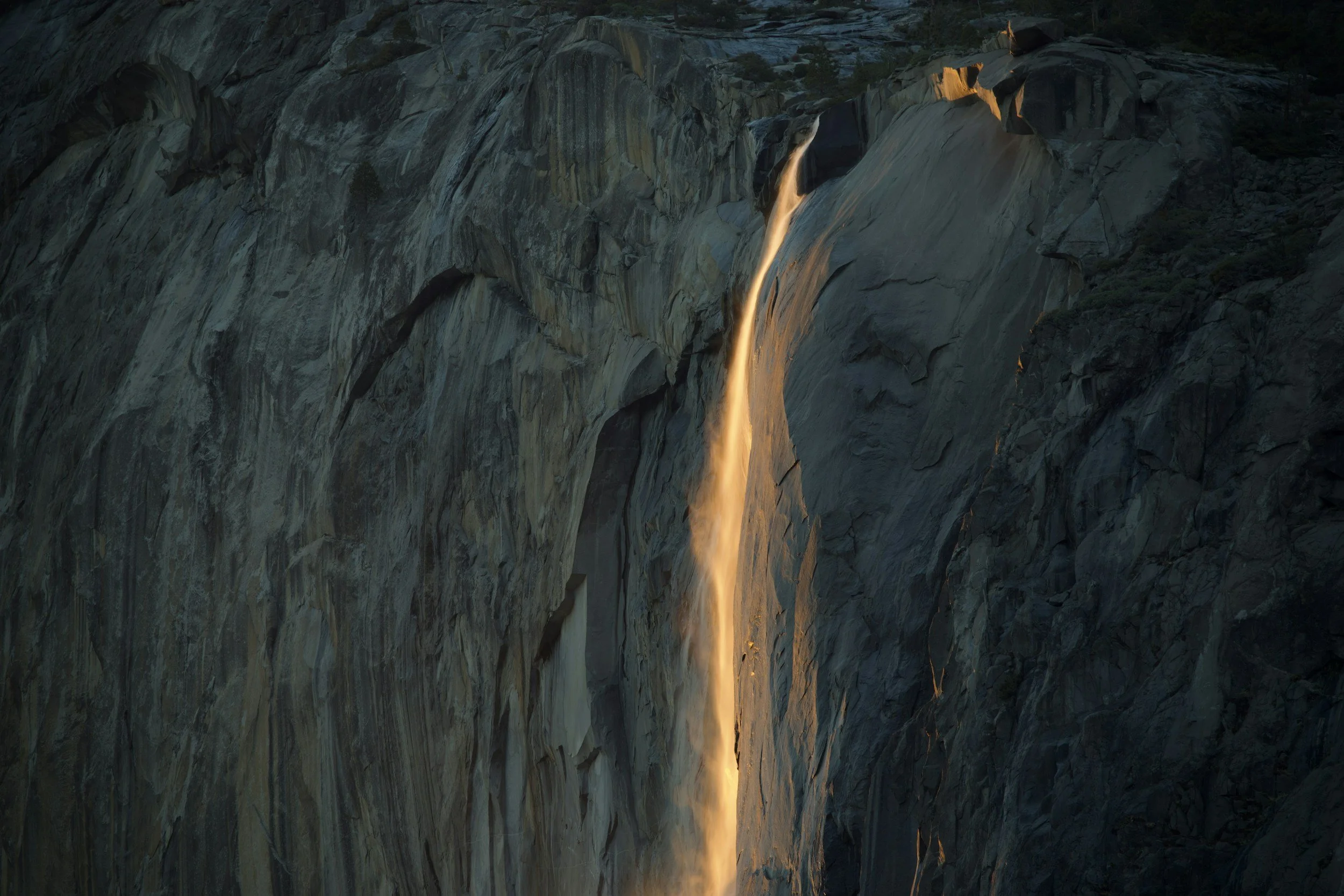 A vertical section of a mountain with a thin waterfall flowing down its face, illuminated by sunlight.
