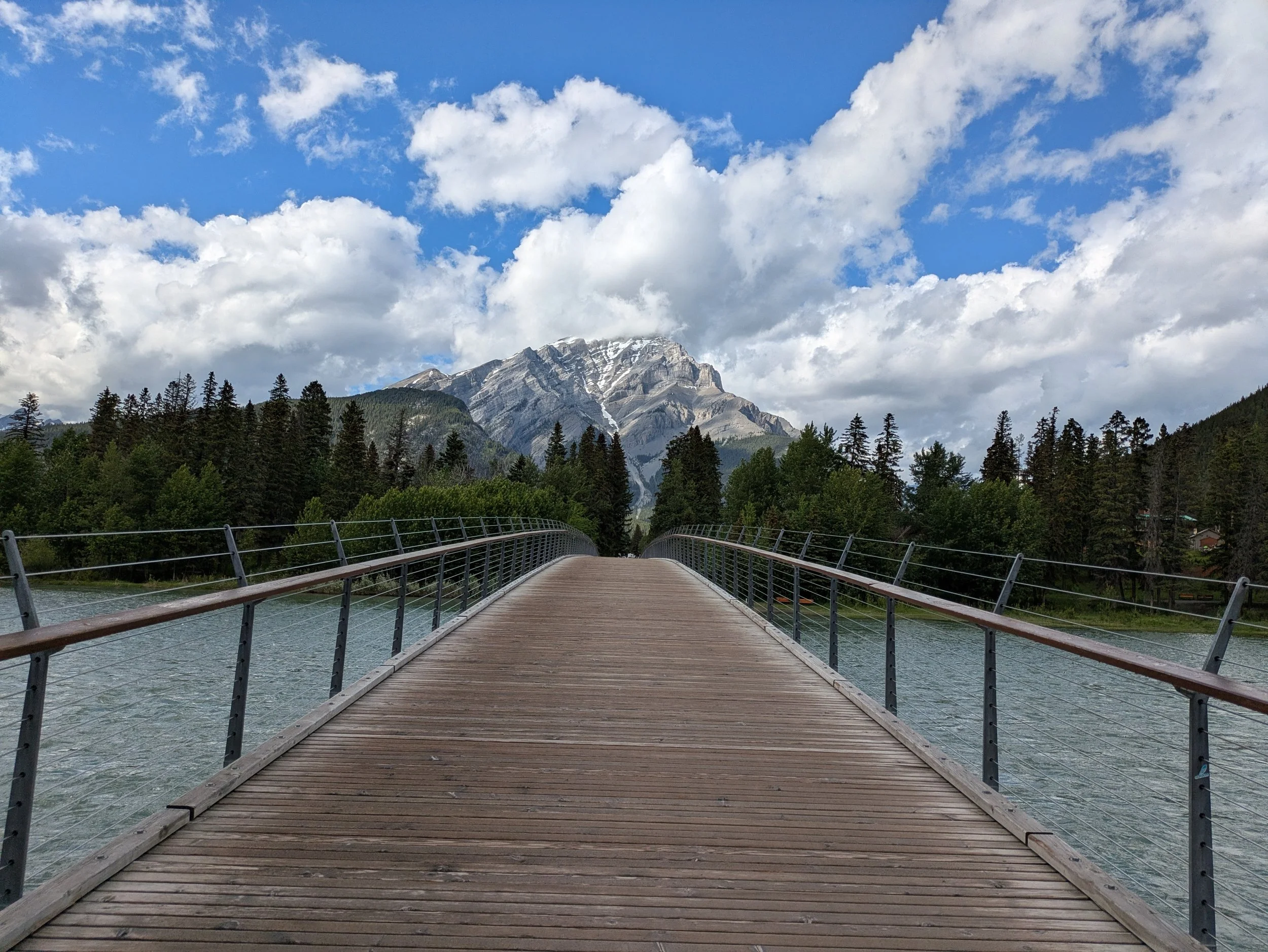 Wooden pedestrian bridge across a river with mountain range in the distance, trees along the riverbank, partly cloudy sky.