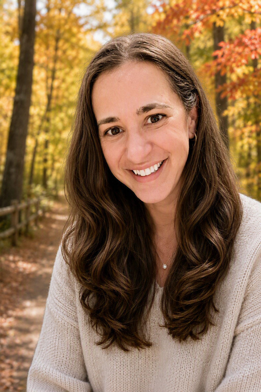 A woman smiling outdoors in a forest with fall foliage, wearing a beige sweater and a necklace.
