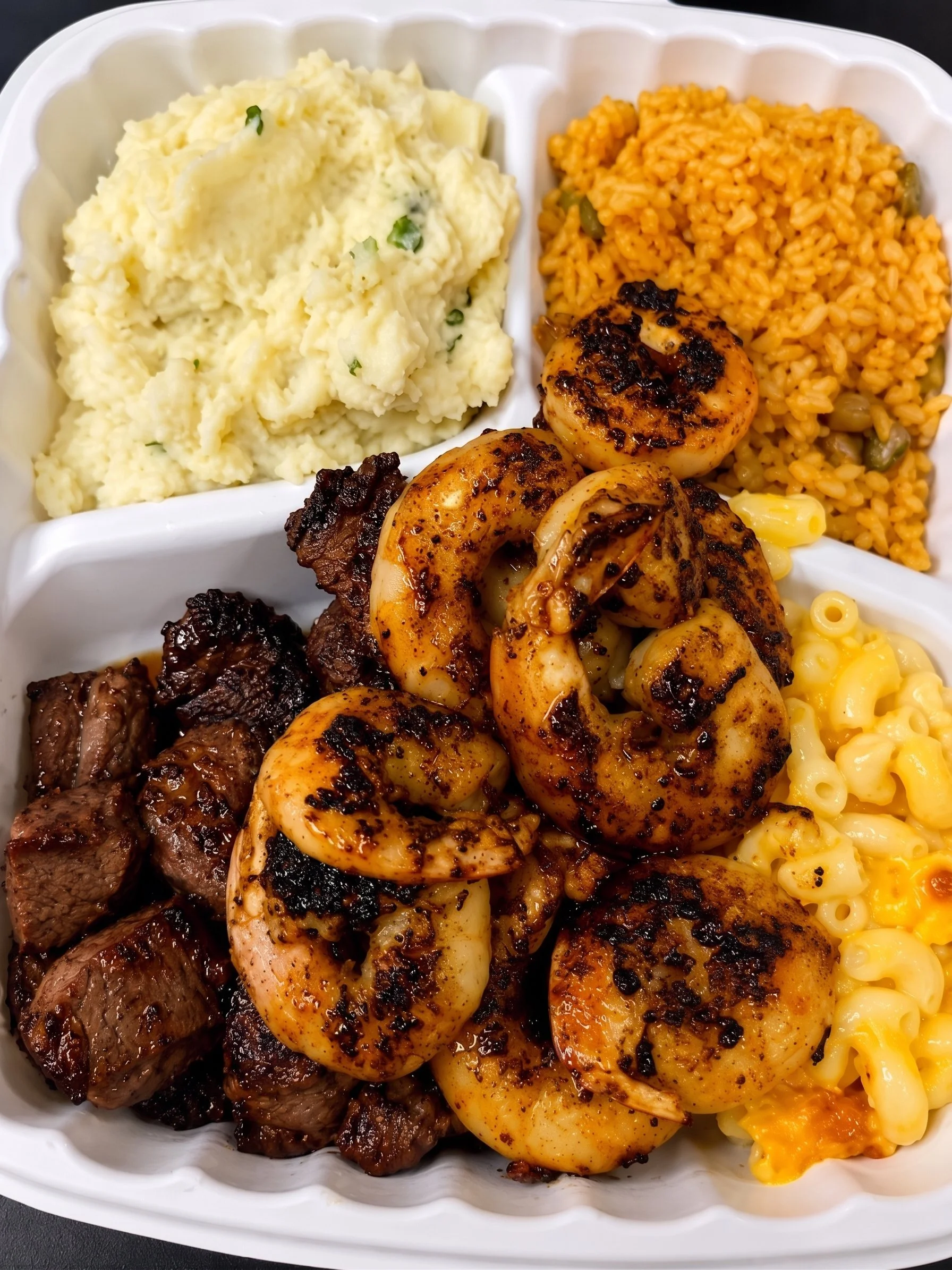 A meal in a white divided tray with mashed potatoes, seasoned rice, grilled shrimp, beef chunks, and macaroni and cheese.