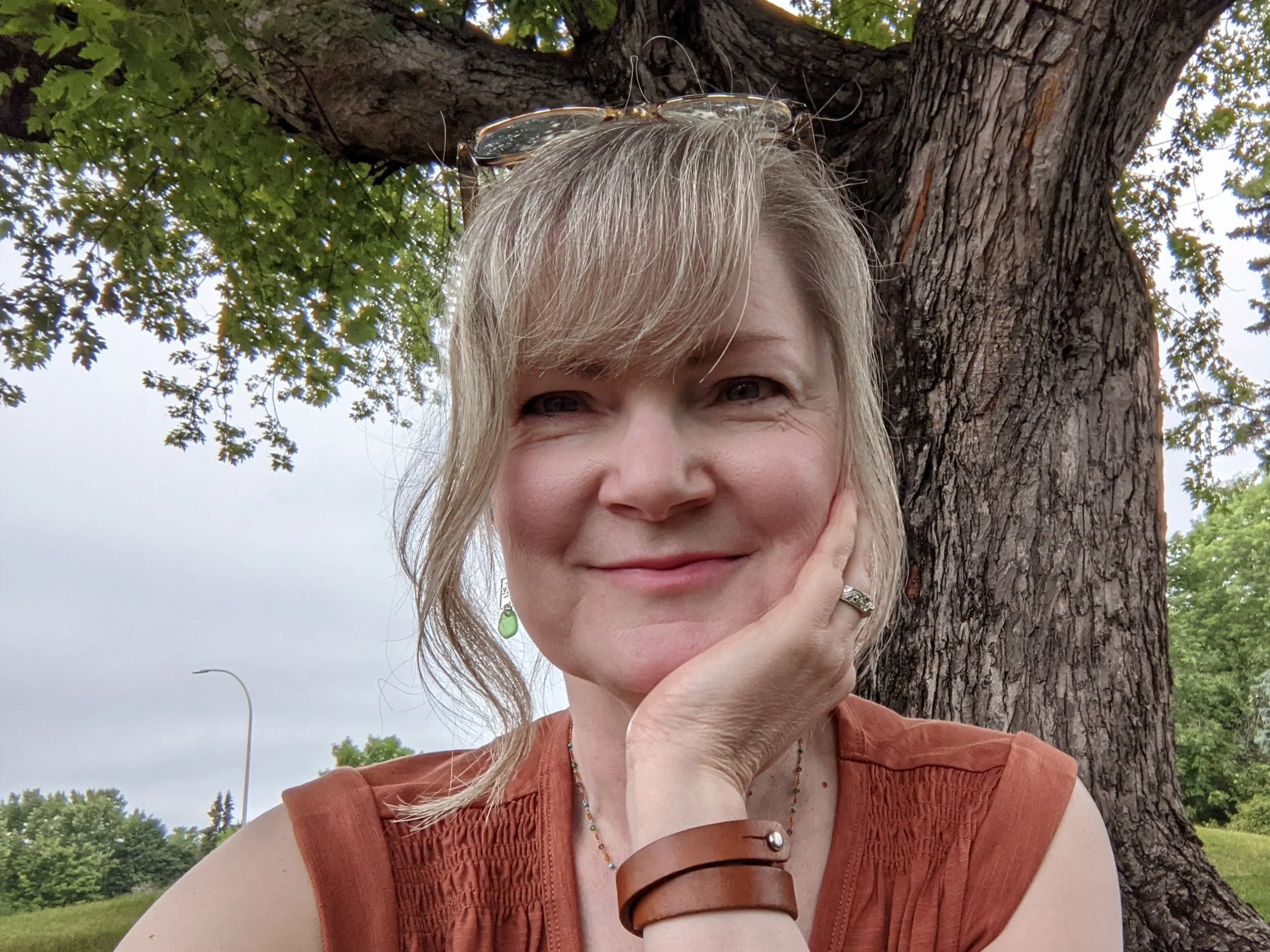 A woman stands outdoors in front of a tall tree with green leaves, smiling and resting her face on her hand. She has short blonde hair, glasses on her head, and wears a rust-colored sleeveless top and jewelry. The background includes a cloudy sky and a lush, green landscape.