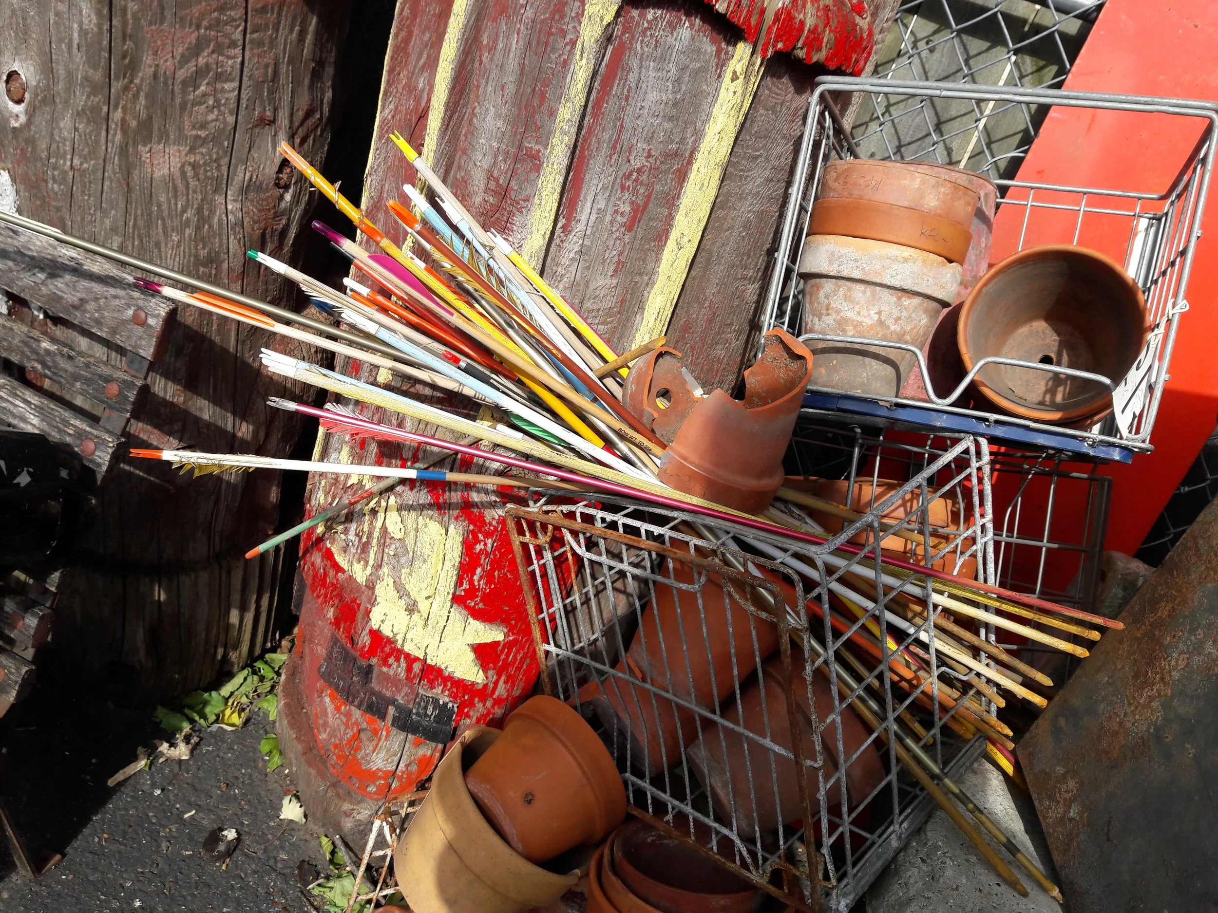 Collection of colorful brooms with long sticks, empty flower pots, and a wire shopping cart on a weathered red wooden surface outdoors.