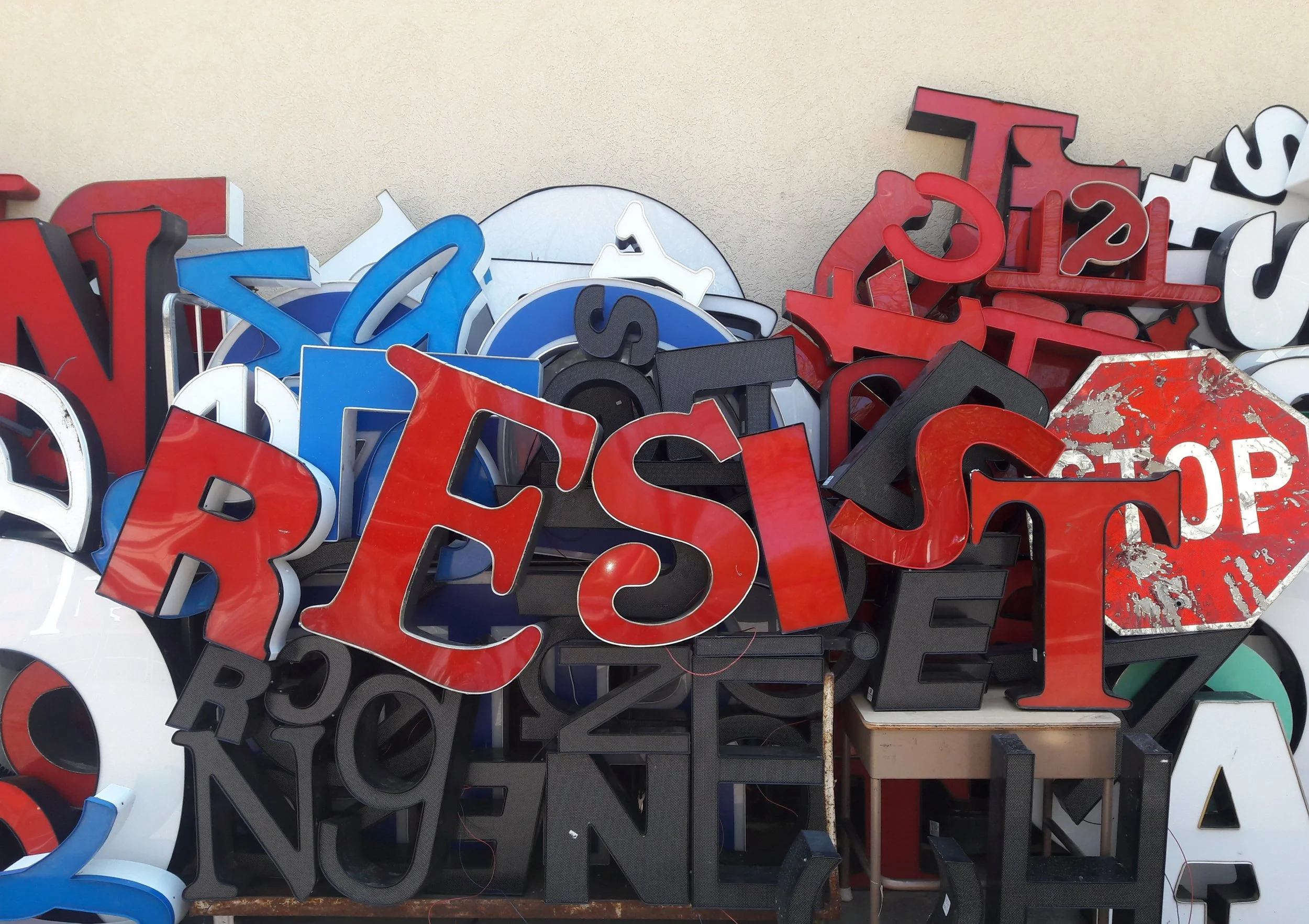 A pile of brightly colored, vintage neon signs with various letters and numbers, some showing weathering and rust, stacked against a plain wall.