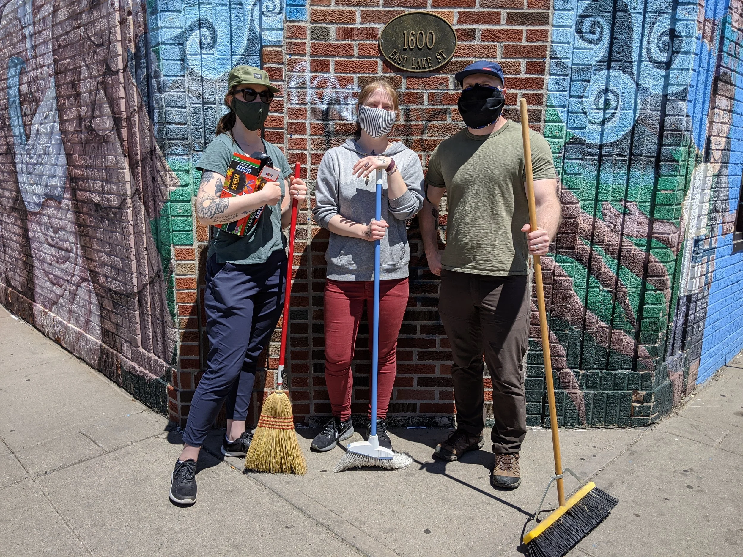 Three people wearing masks and holding cleaning tools, standing in front of a brick wall with a street address plaque that reads 1600 East Lake Street.