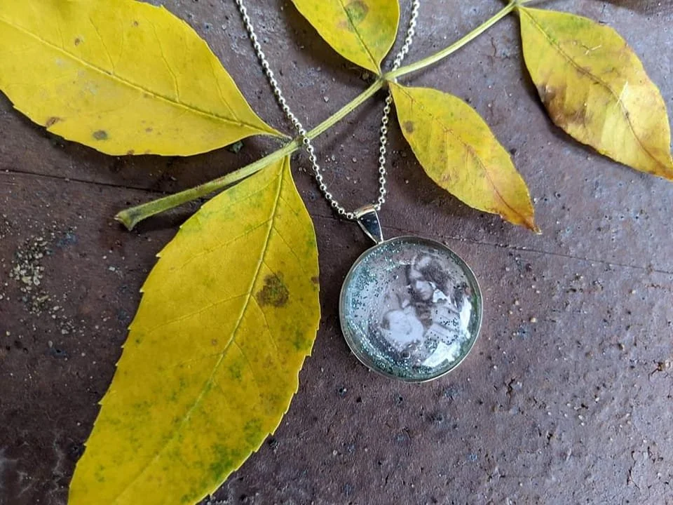 A silver necklace with a round pendant featuring a black and white photo of two people, laid on a wooden surface with yellow leaves.