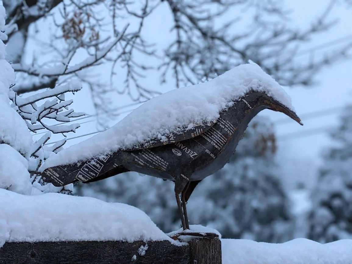 A metal bird sculpture covered with snow, perched on a wooden surface with snow, with snow-covered tree branches in the background.