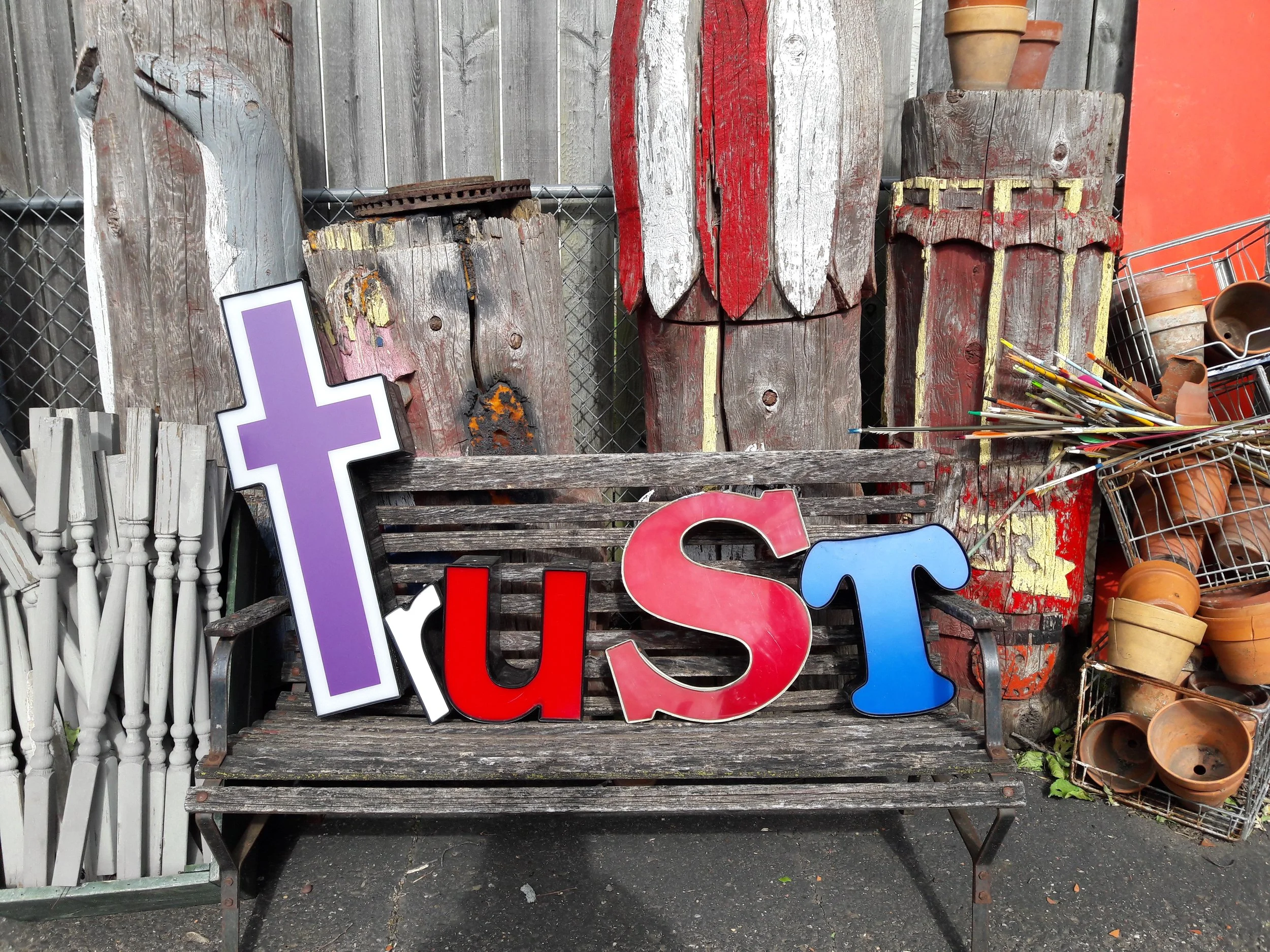 A rustic wooden outdoor display shelf holding potted plants, some decorative wooden pieces painted in red, white and yellow, and colorful plastic letters spelling 'HIS'.