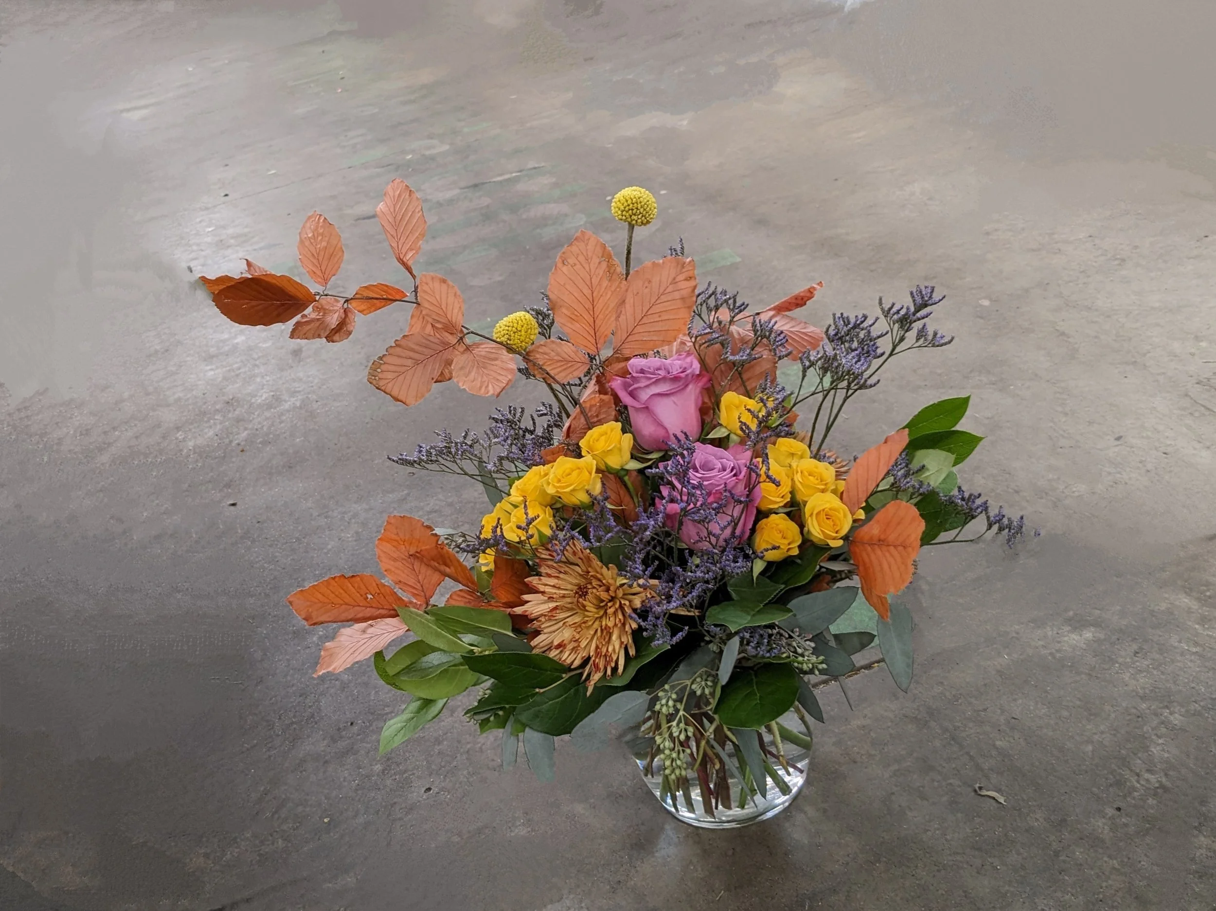 A floral bouquet arranged in a glass vase, featuring pink roses, yellow flowers, purple sprigs, and orange-brown leaves, placed on a concrete floor.