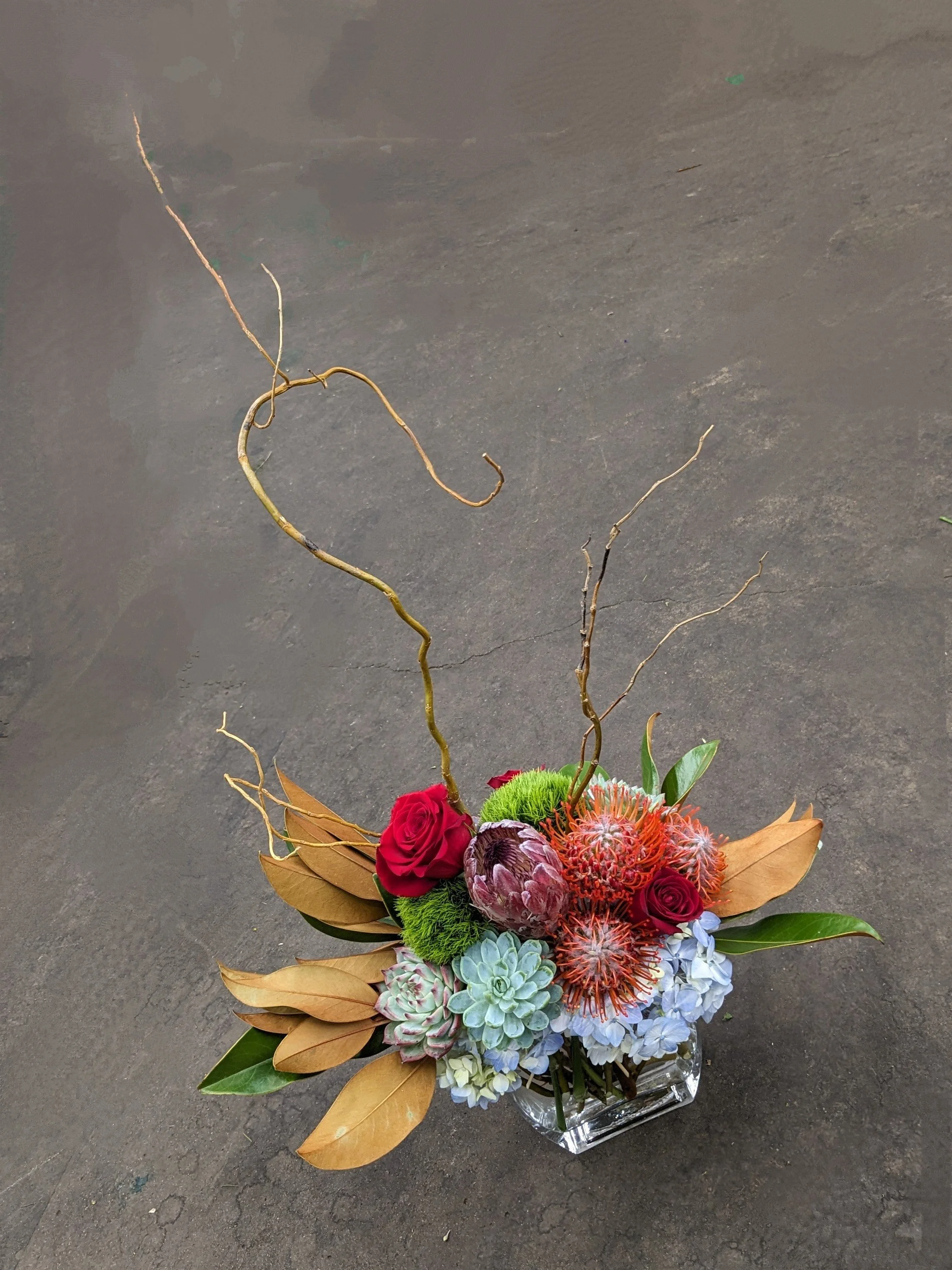 A floral arrangement in a glass vase with various flowers including red roses, blue hydrangeas, succulents, and orange protea, accented with dried leaves and twisted branches, set on a dark surface.
