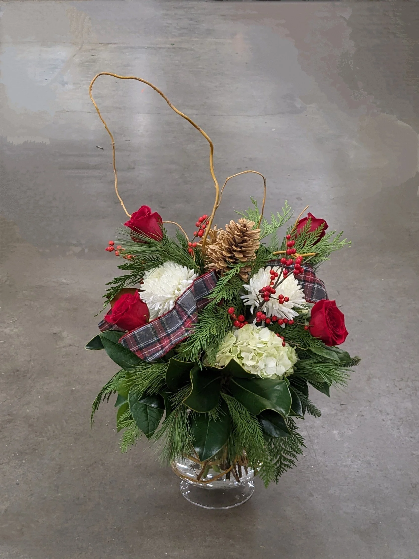 A floral arrangement in a glass vase featuring red roses, white chrysanthemums, pinecones, pine branches, red berries, and a plaid ribbon, set against a concrete floor.
