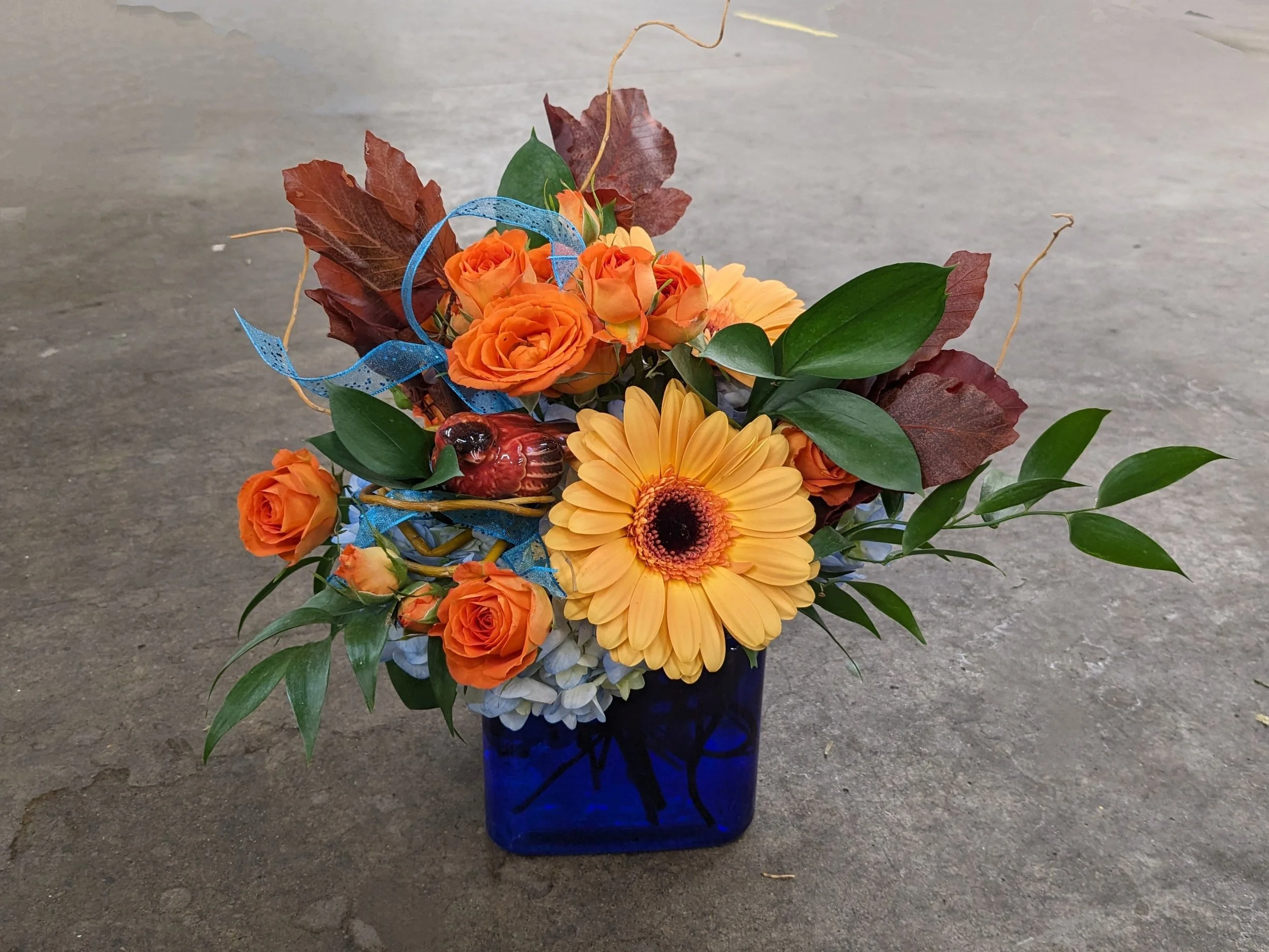 A bouquet of orange roses, yellow gerbera daisies, and various green and brown leaves arranged in a blue glass vase on a concrete or asphalt surface.