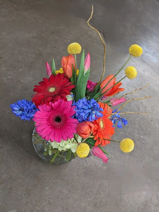 Colorful bouquet of various flowers including pink, red, orange, blue, and yellow blooms in a glass vase on a gray surface.