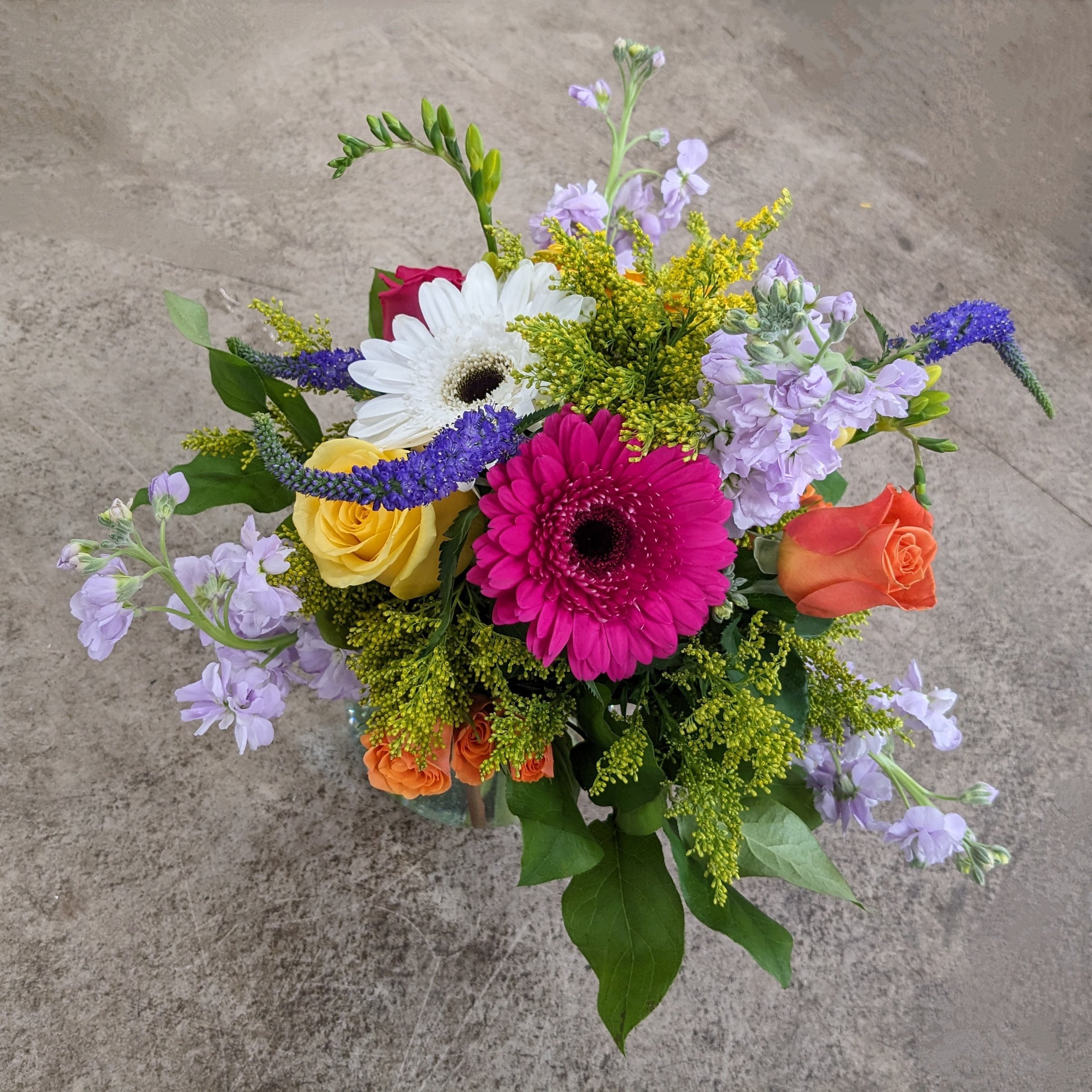A colorful bouquet of flowers including white daisies, pink gerbera daisies, orange roses, and purple statice, set against a neutral background.