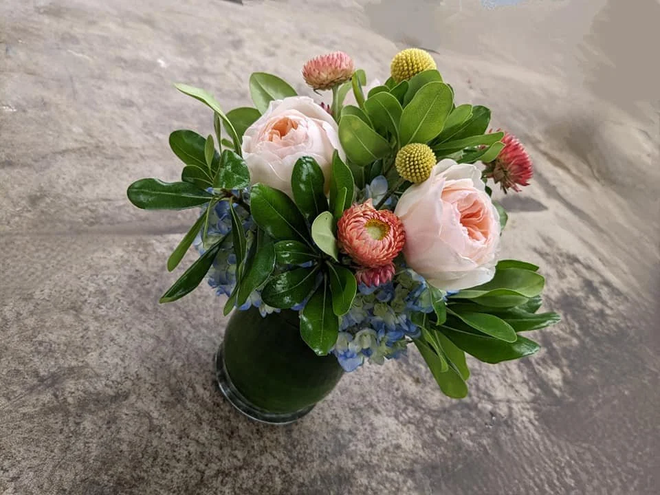 A bouquet of pink roses, pink and yellow button poms, blue hydrangeas, and green leaves in a dark green glass vase on a gray stone surface.