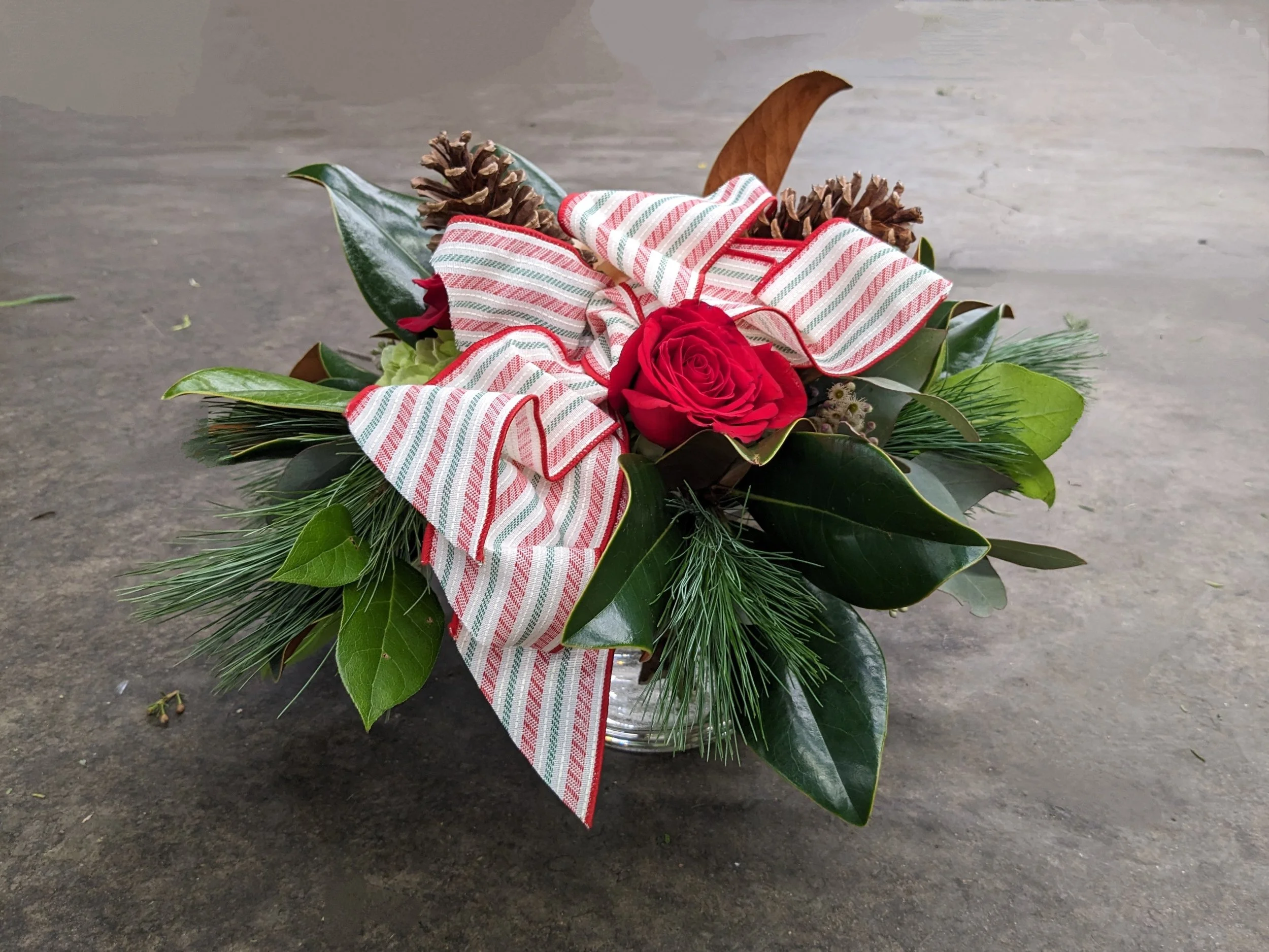 A Christmas floral arrangement in a glass container, decorated with a red and white striped ribbon, featuring a red rose, pinecones, and green leaves.